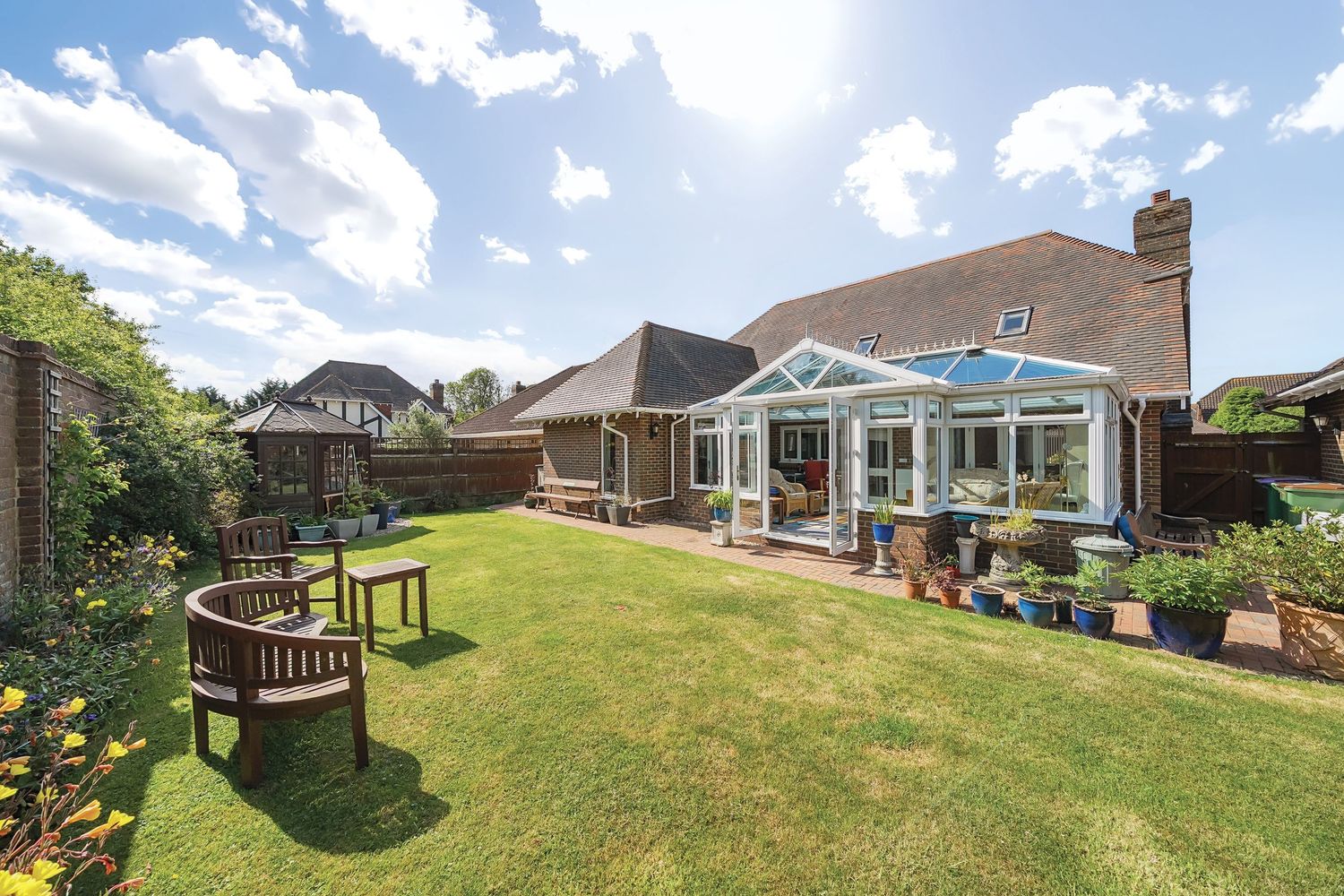 Beautiful residential garden with lush green lawn, wooden benches, and large conservatory extension on brick house.
