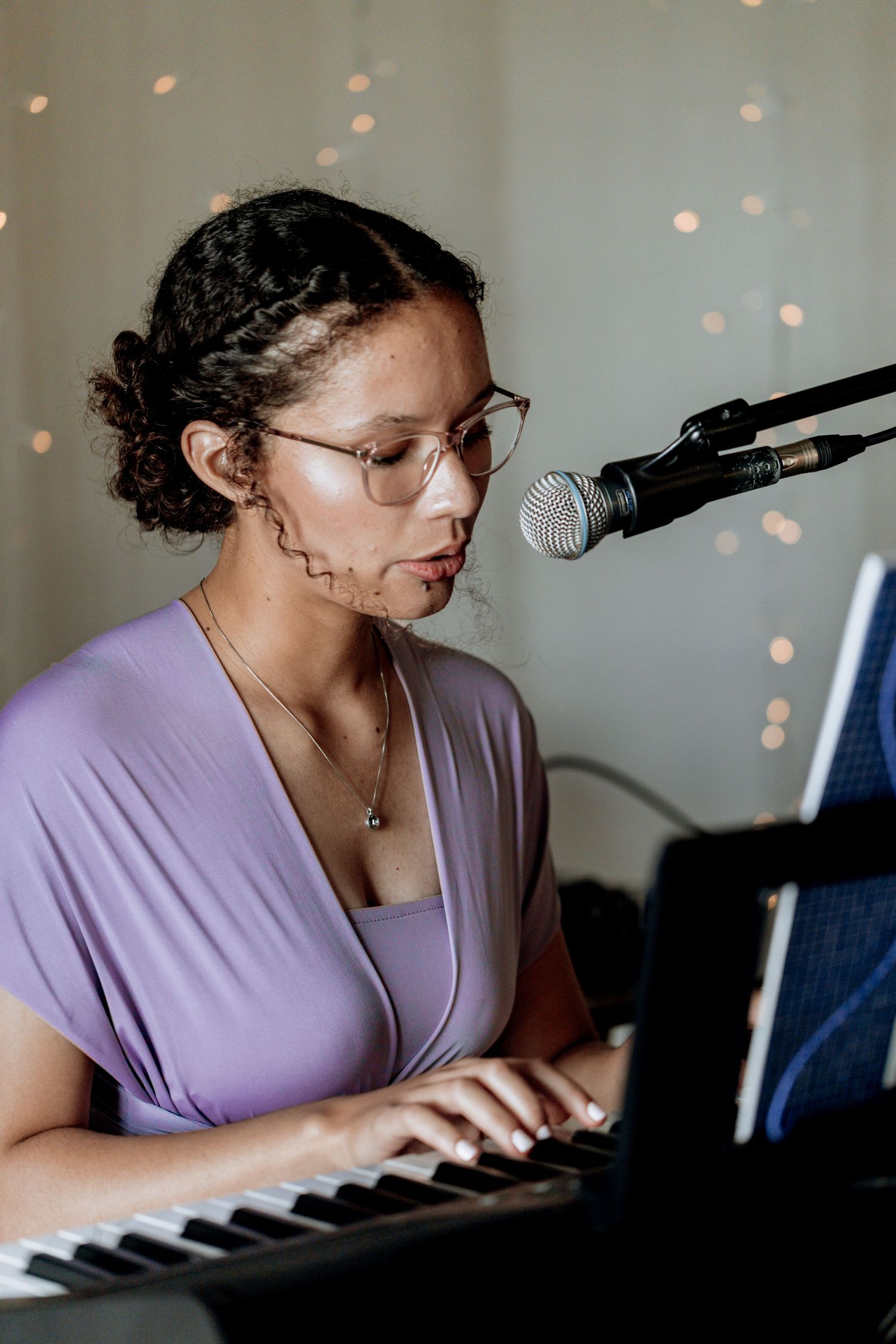 Musician in a purple top plays keyboard while singing into a microphone with twinkle lights in the background.