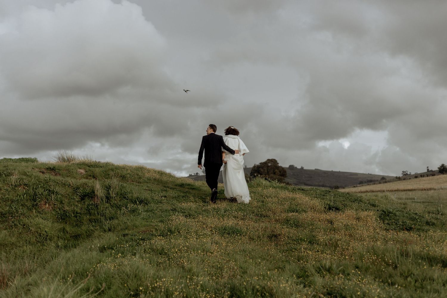 A couple walks together on a scenic hilltop overlooking green rolling hills under dramatic cloudy skies.