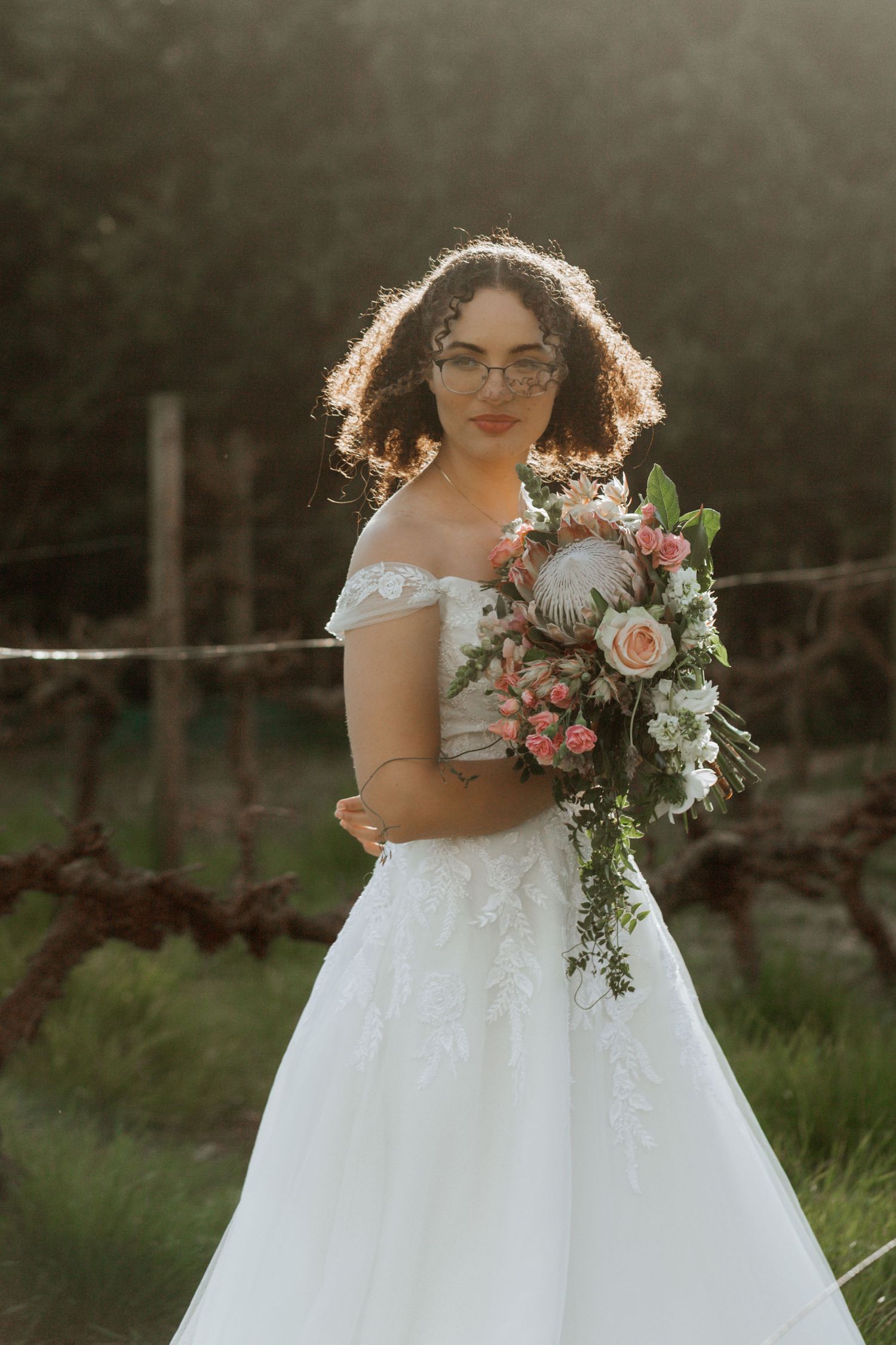 Bride in a white off-shoulder wedding gown holds a romantic pink and white floral bouquet in an outdoor garden setting.