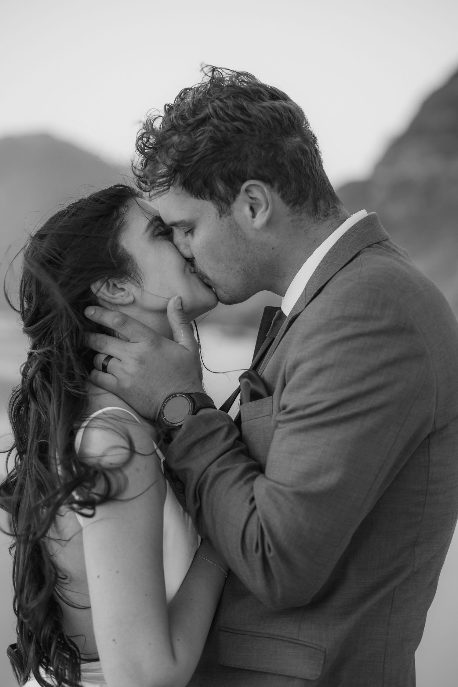 Black and white photo of a couple sharing a romantic kiss, groom in suit holding bride's face tenderly.