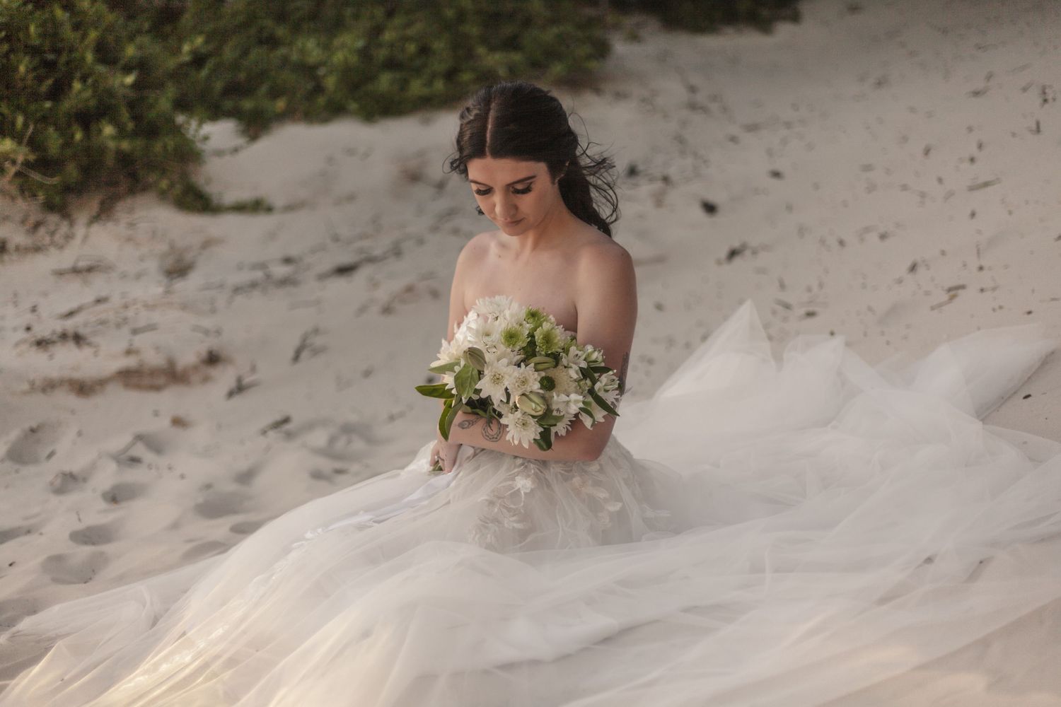 Bride in off-shoulder tulle gown holding white floral bouquet on sandy beach.