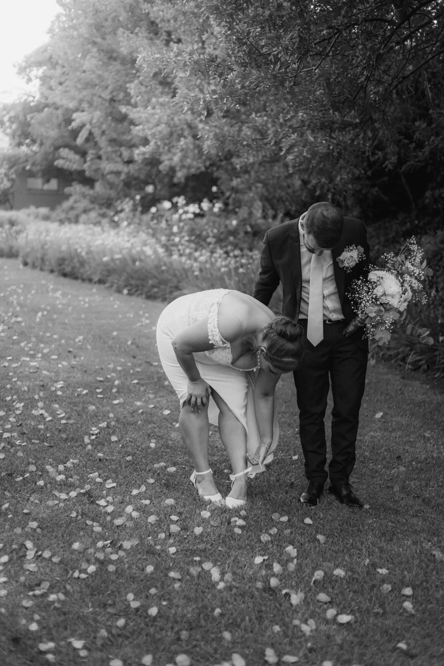 Black and white photo of a bride adjusting her heel while groom watches in a garden scattered with flower petals.