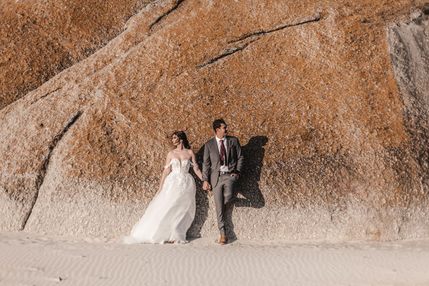 Bride and groom hold hands on sandy beach beside a large textured rock formation during their outdoor wedding photo session.