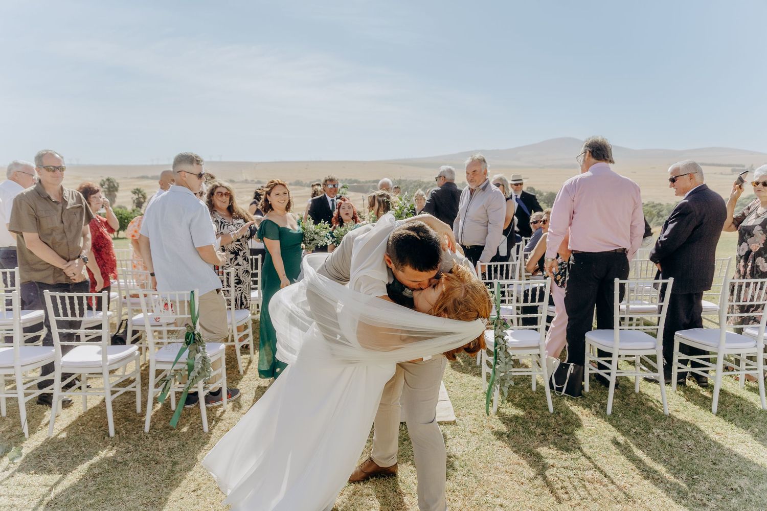 Bride and groom share a romantic dip kiss at outdoor wedding ceremony with white chairs and guests.