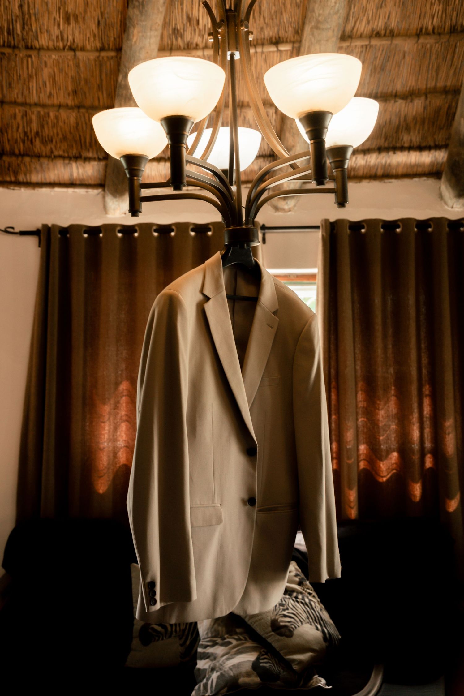 A light gray suit jacket hanging on a chandelier with glowing bulbs, brown curtains and thatched ceiling in background.