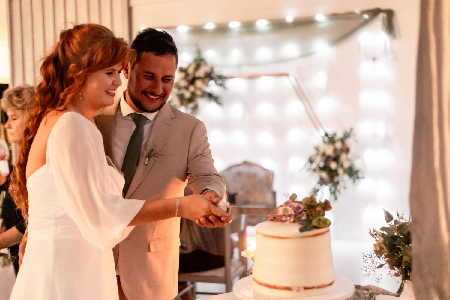 Bride and groom smile while cutting white wedding cake under string lights at elegant tent reception.
