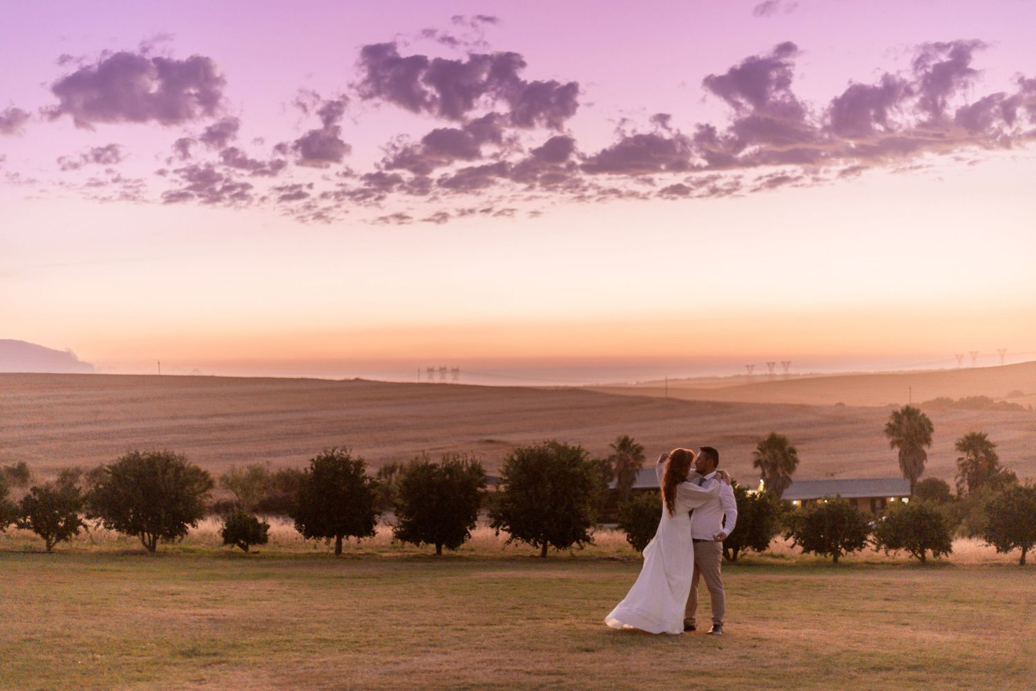 Couple embracing at sunset on a grassy hill with golden fields and purple sky backdrop.