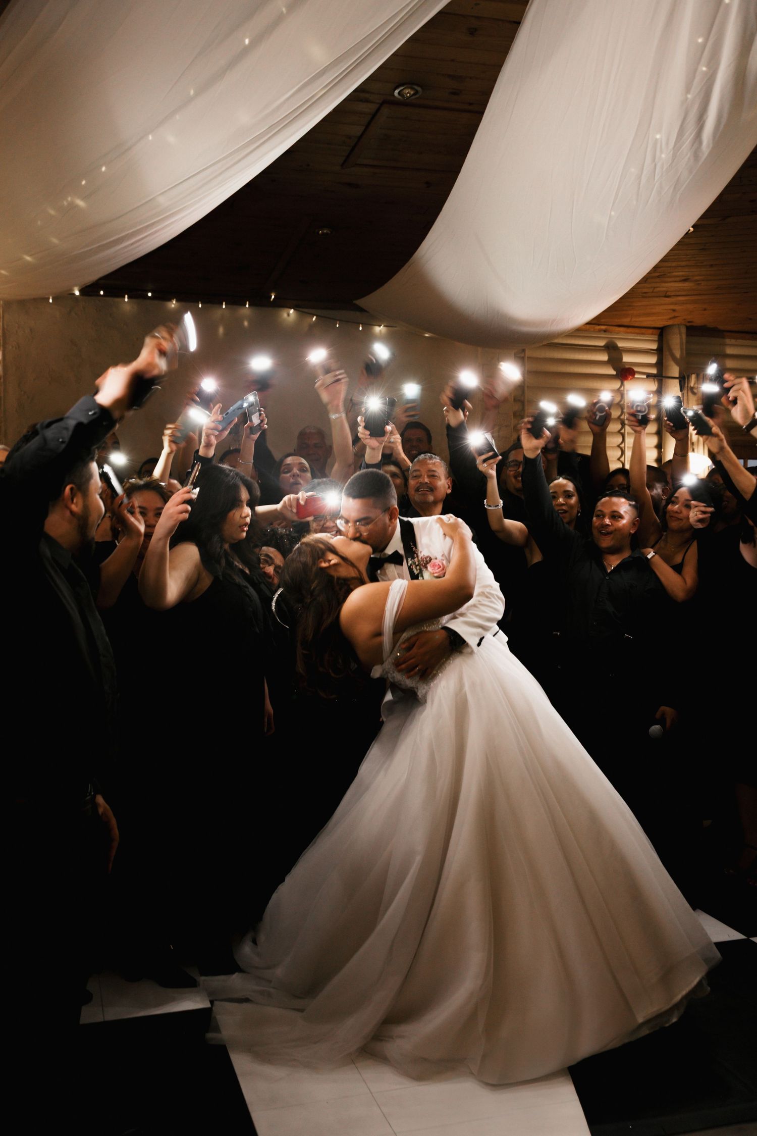 Bride and groom share a romantic dip kiss on the dance floor surrounded by wedding guests holding up glowing phone lights.