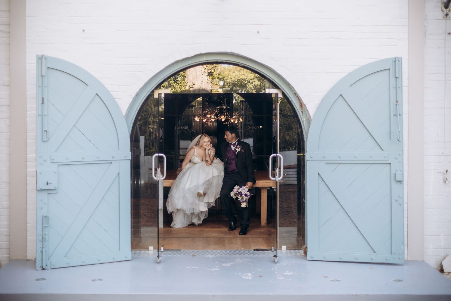 Bride and groom exit through arched double doors of a white chapel with light blue barn doors at their wedding ceremony.