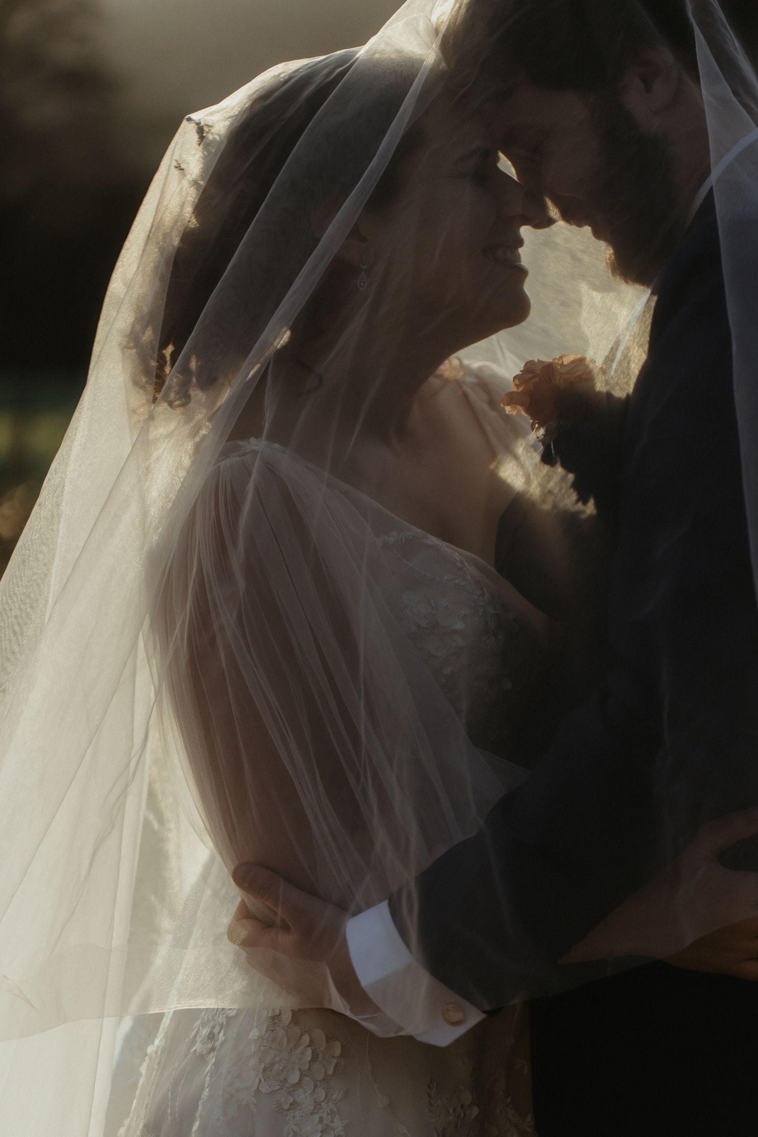 A romantic silhouette of a bride and groom kissing under a flowing white veil during their wedding ceremony.