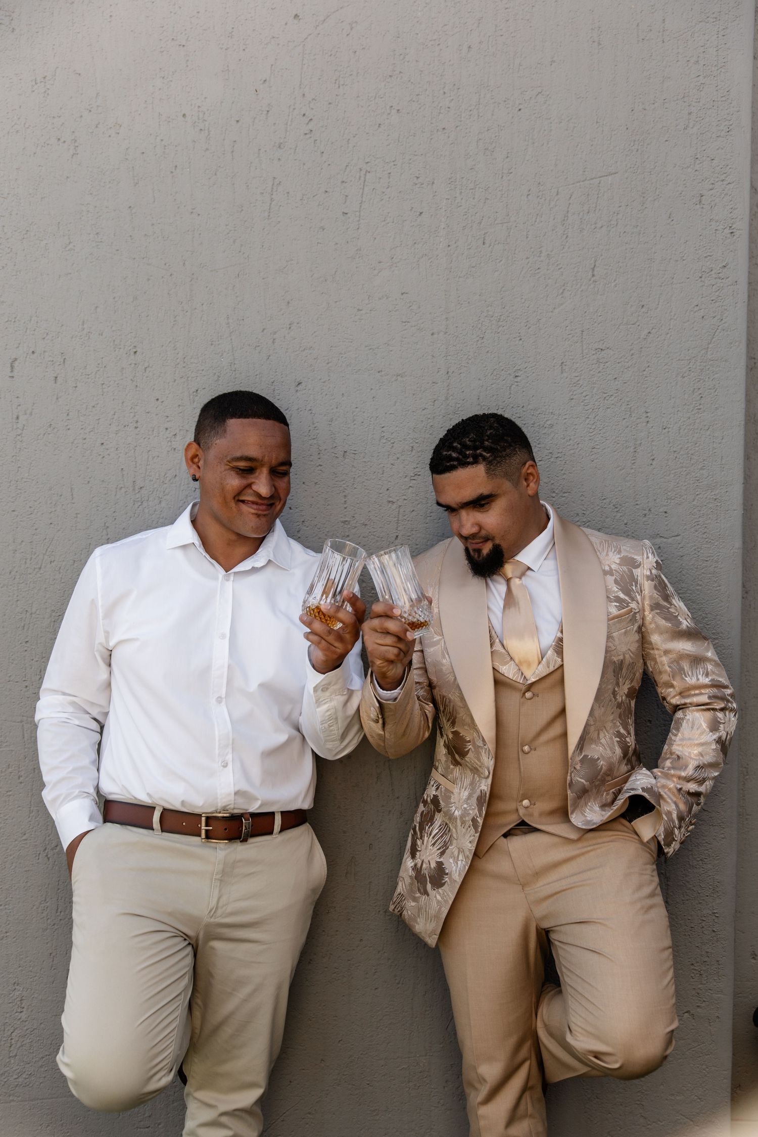 Two people in coordinating beige and white outfits lean against a gray wall while laughing together.