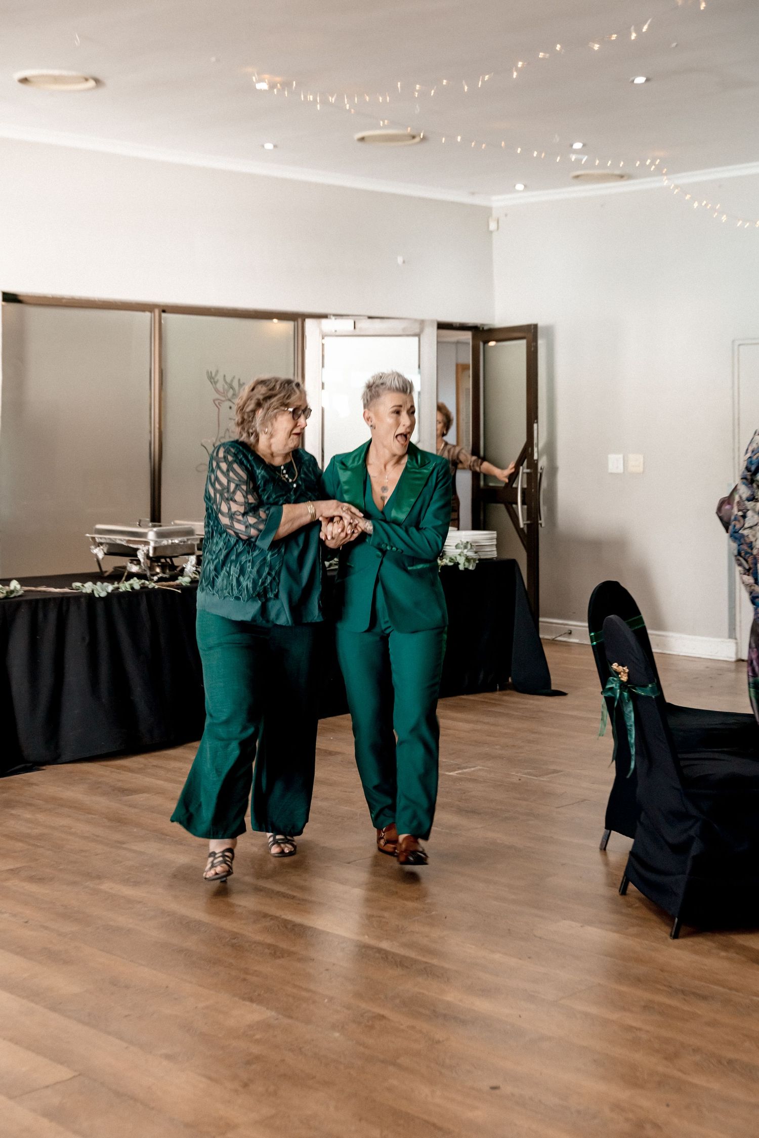Wedding guests in matching emerald green outfits dancing together in a reception venue with string lights.