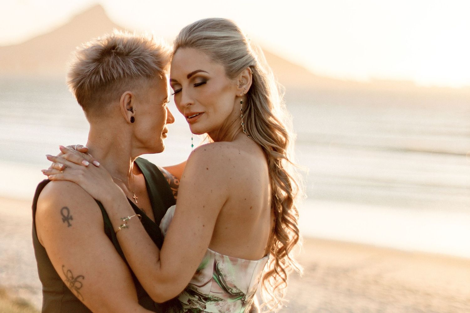 A romantic couple shares an intimate moment on a beach at sunset with mountains in the background.