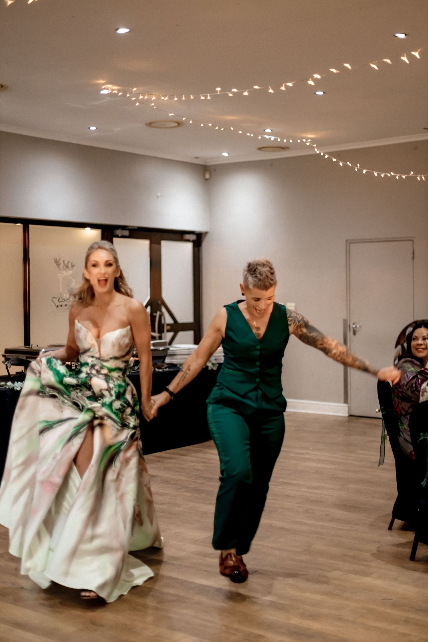 A group of wedding guests dance together on a wooden floor under string lights during a reception celebration.