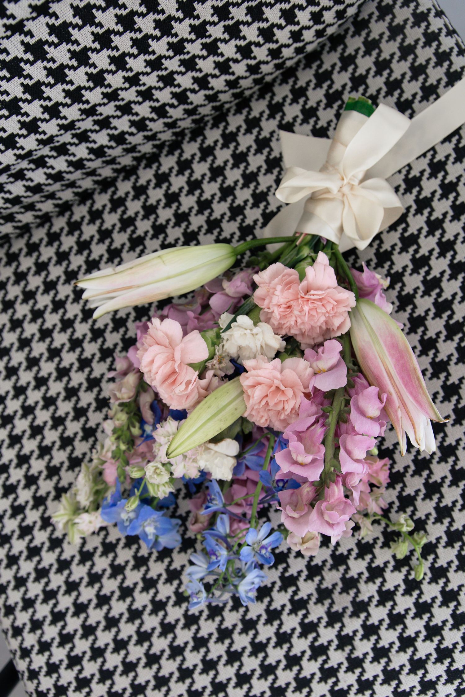 A pink and blue floral bouquet featuring carnations and delphinium arranged on a black and white houndstooth pattern background.