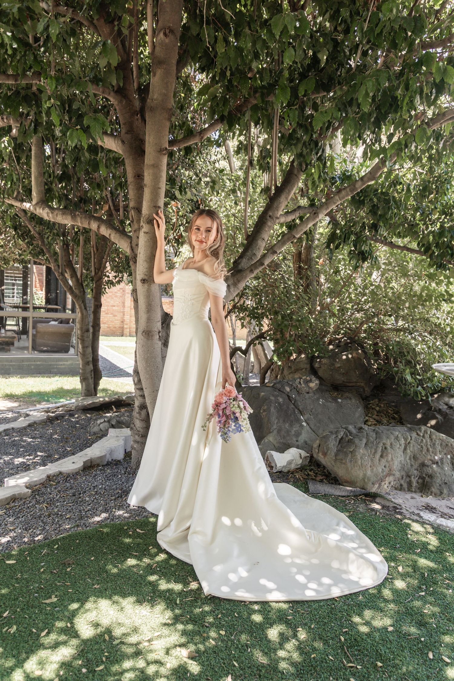 Elegant wedding dress with off-shoulder design and long train photographed in dappled sunlight under trees in garden setting.