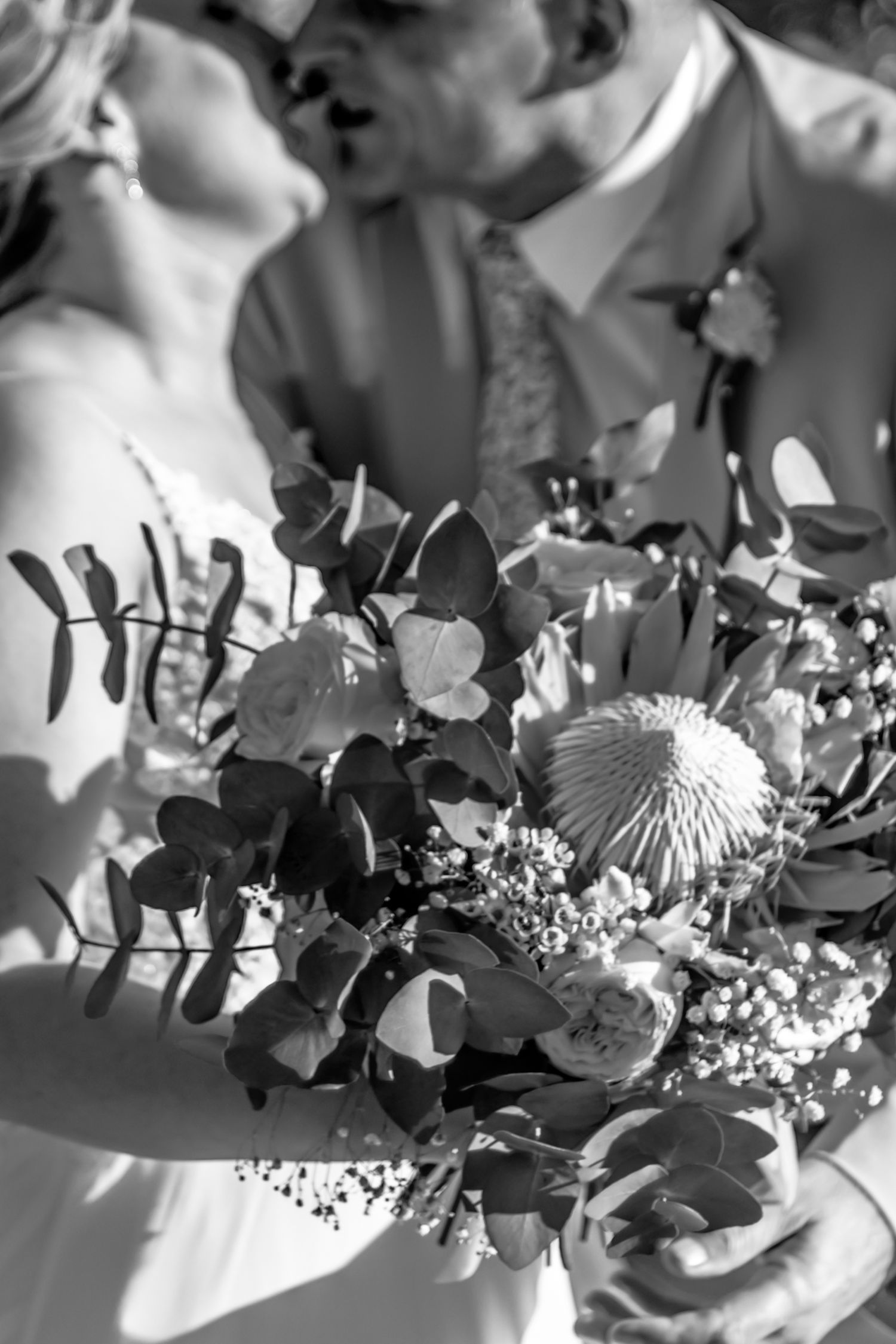 A romantic black and white wedding photo featuring a bouquet of eucalyptus and protea flowers in the foreground.
