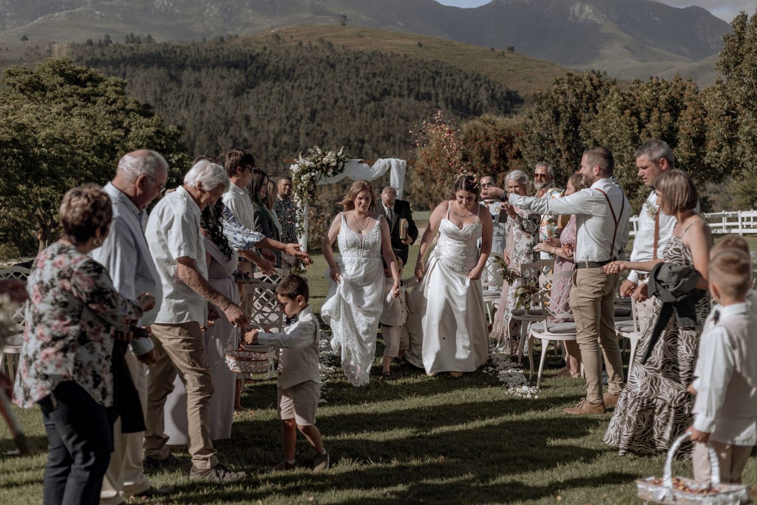 Wedding guests gathered outdoors in white attire celebrating in front of scenic mountain landscape backdrop.