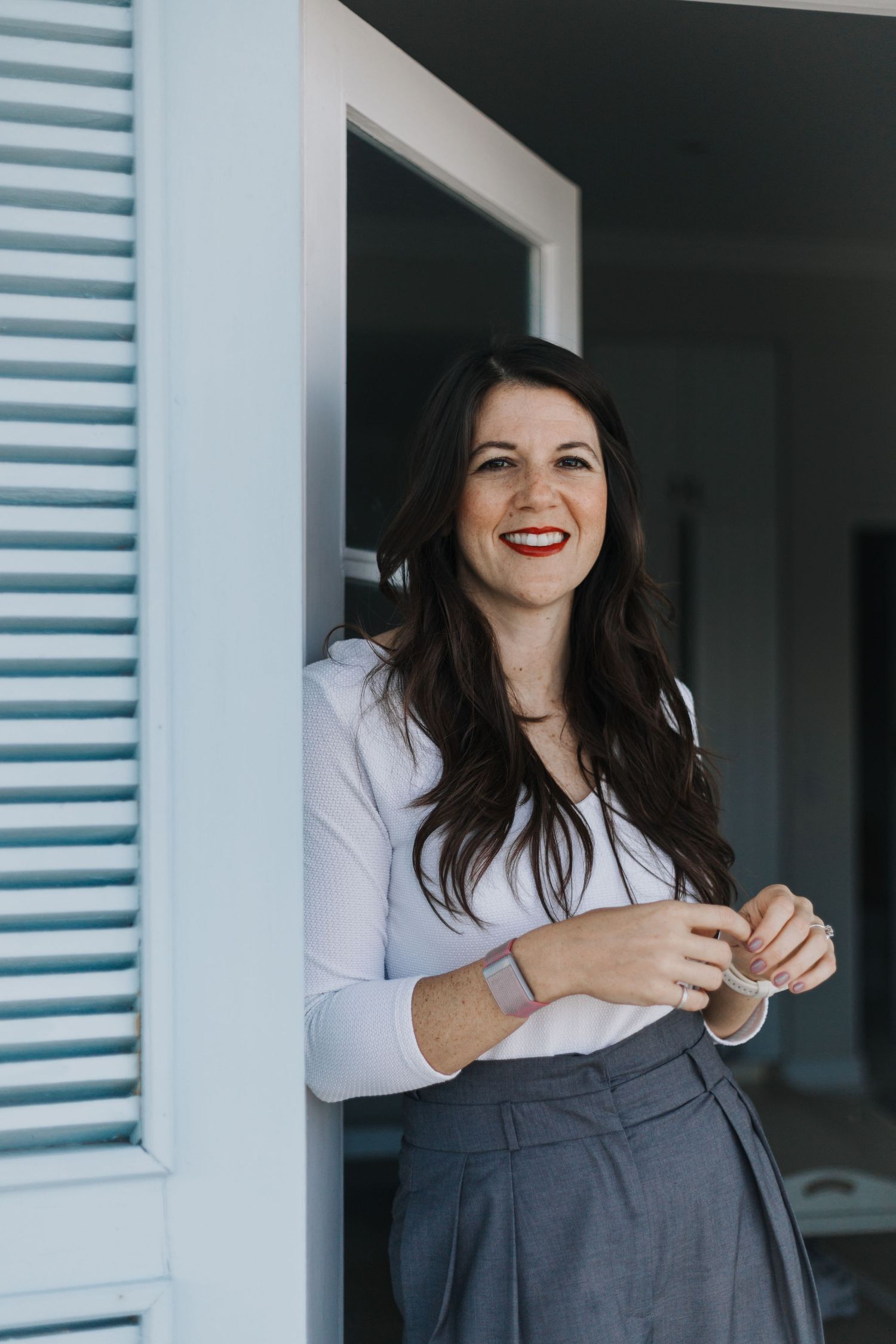 A woman in a dark vest and white top smiles while posing near a blue-shuttered building.