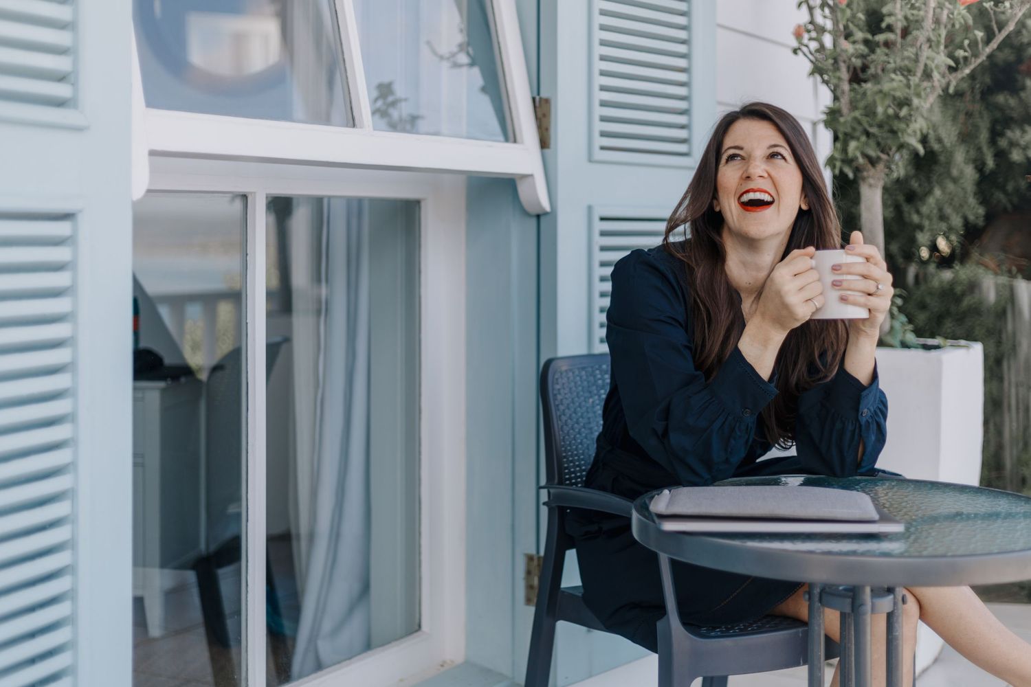 Someone in a navy sweater laughs while sitting on an outdoor chair near blue shuttered windows.