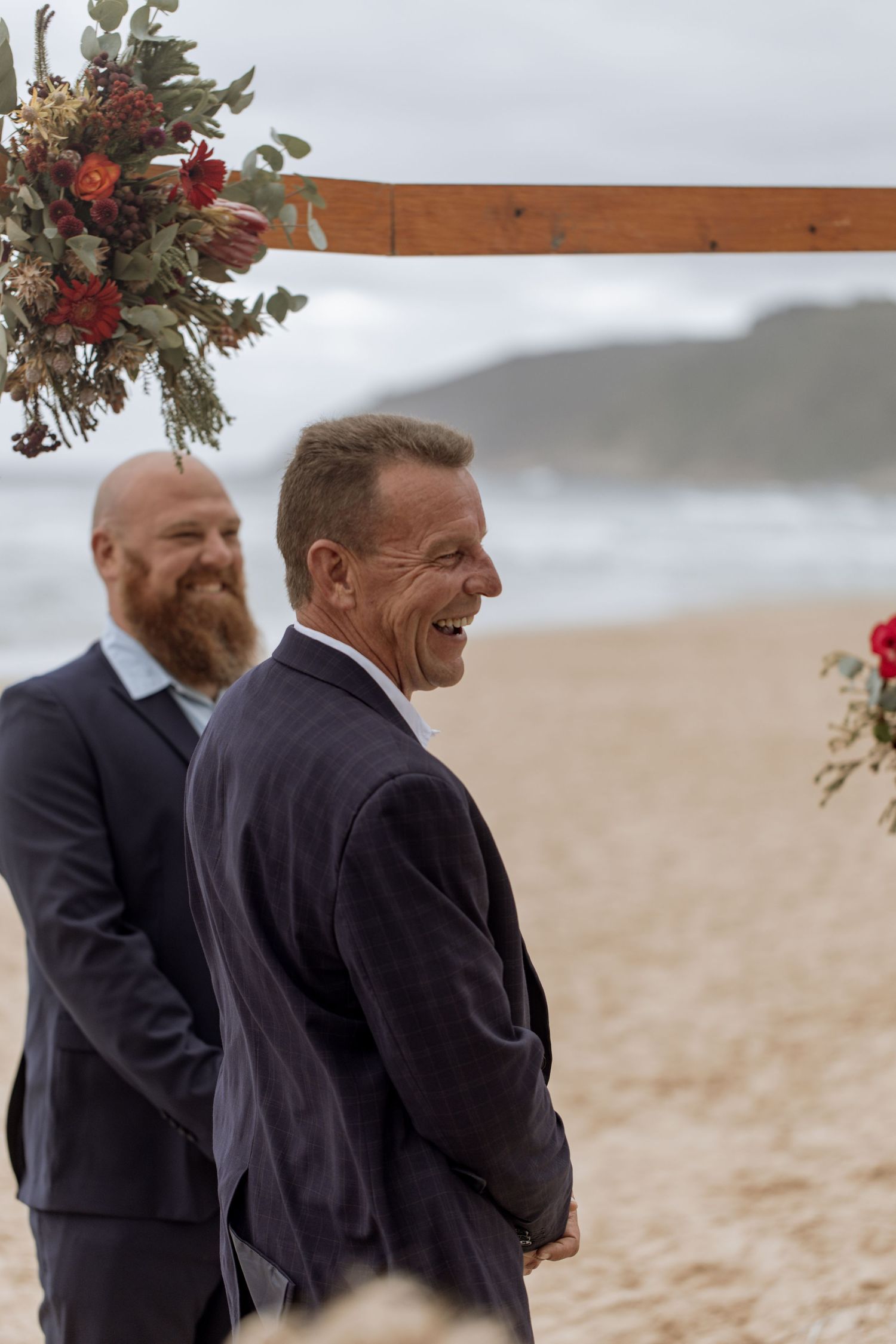 Men in formal suits stand under a wooden arch decorated with floral arrangements at a beach wedding ceremony.