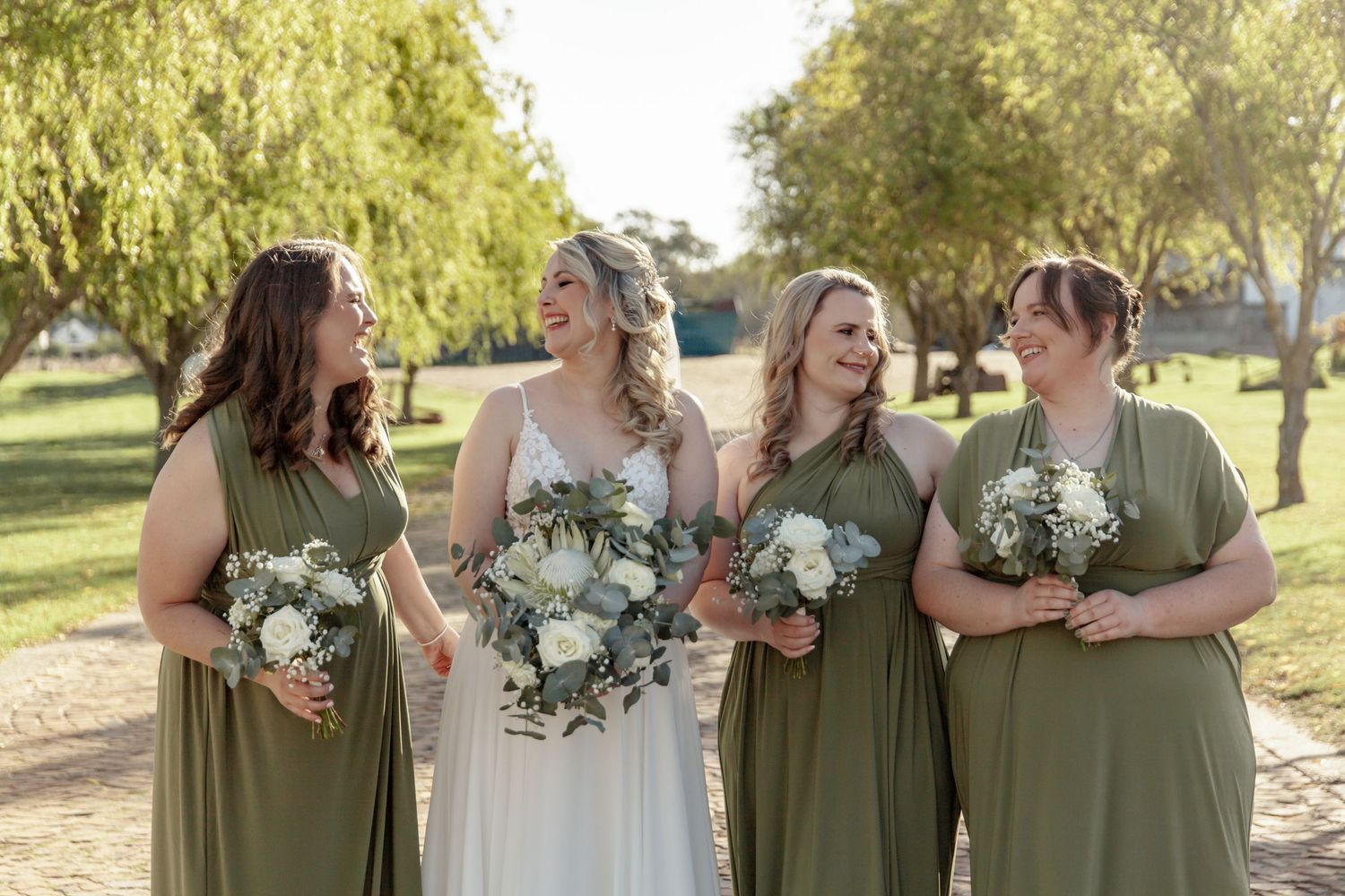 A bridal party poses together outdoors wearing sage green dresses and holding white bouquets.