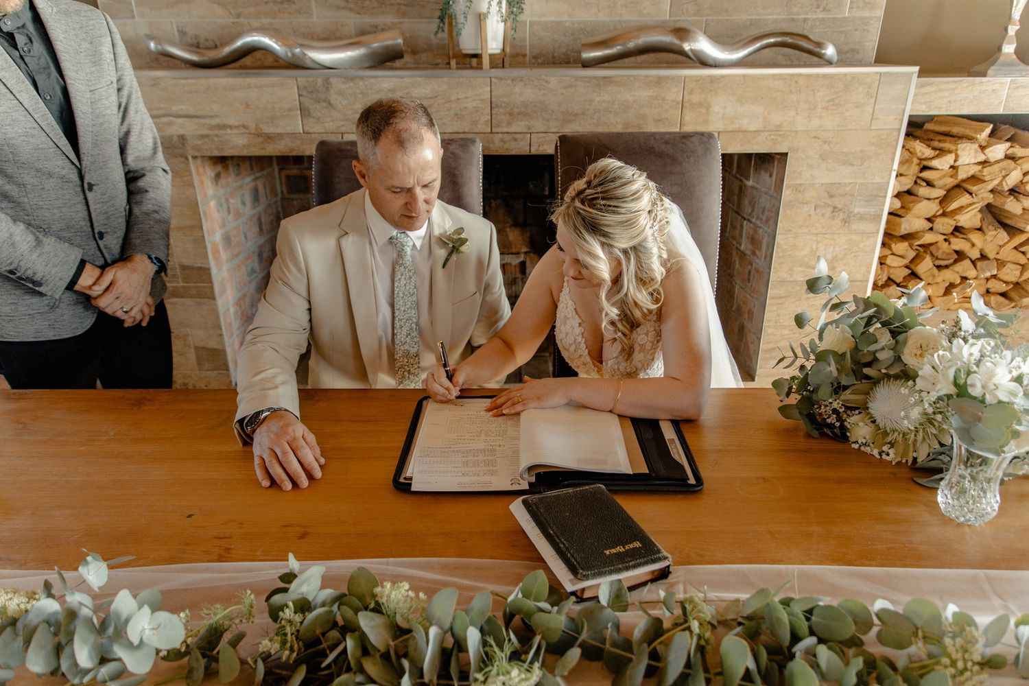 Couple signing marriage documents at wooden table decorated with eucalyptus greenery and white floral arrangements.