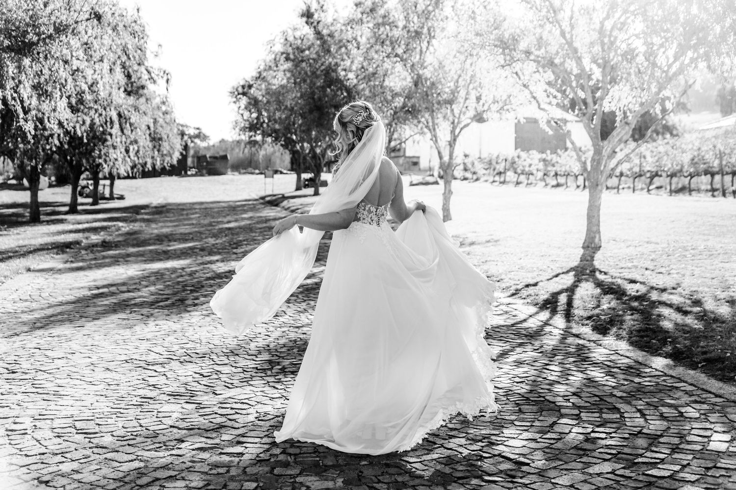 Black and white photo of a bride in a flowing wedding dress twirling outdoors among trees in a garden setting.