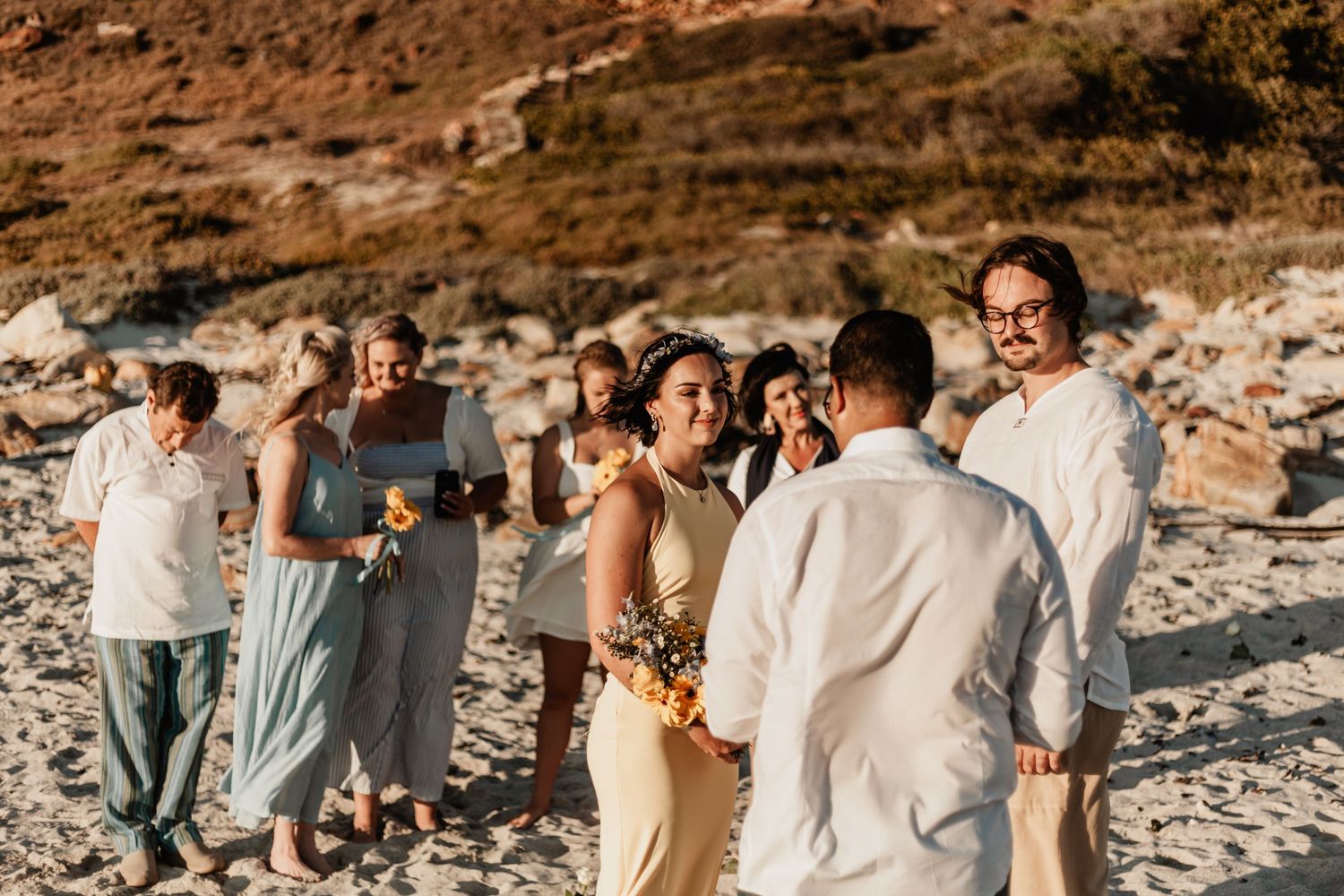 Wedding party stands together on rocky beach at sunset wearing light colored casual attire.