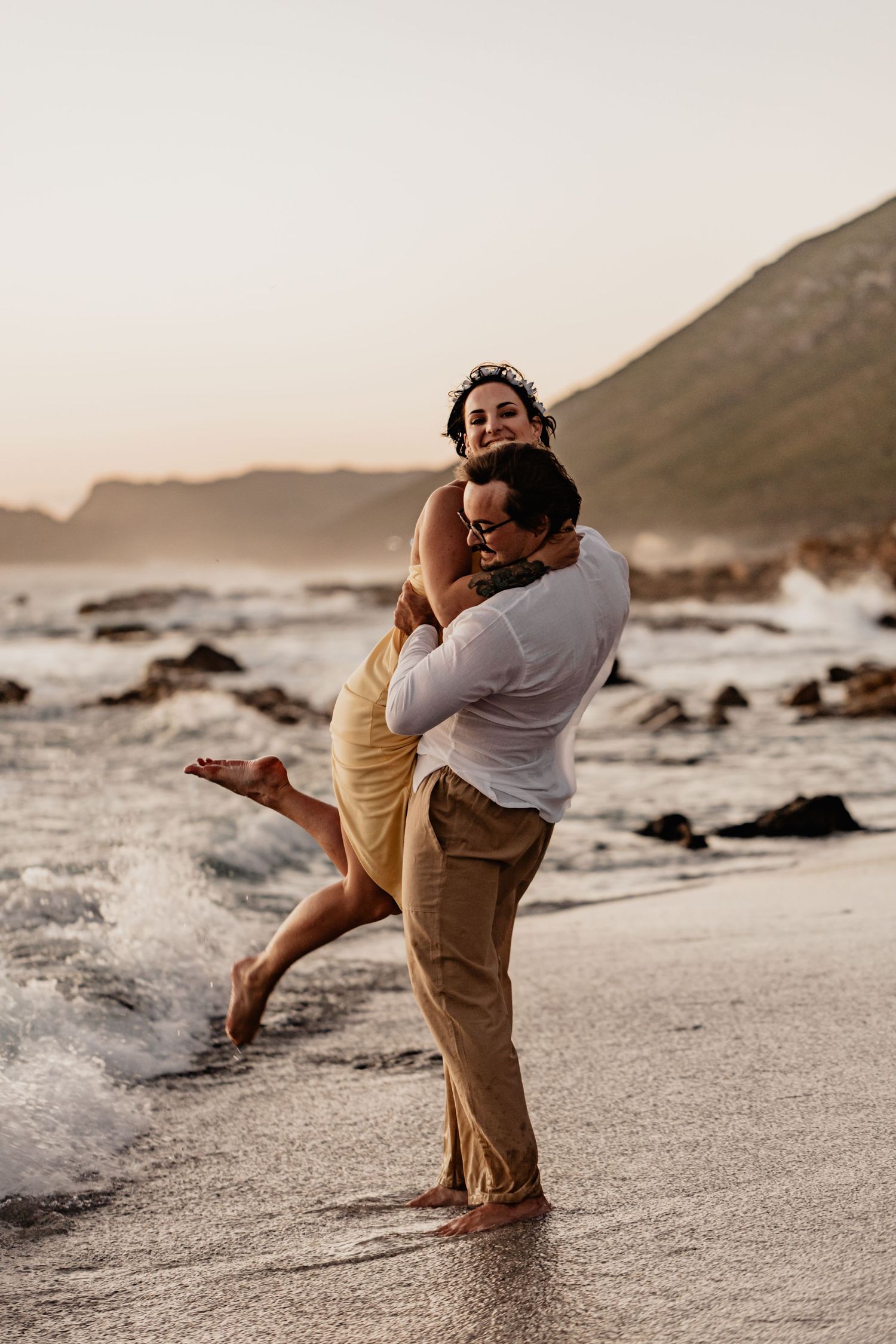 A couple shares an intimate dance embrace on a scenic beach at sunset with mountains and coastline in the background.