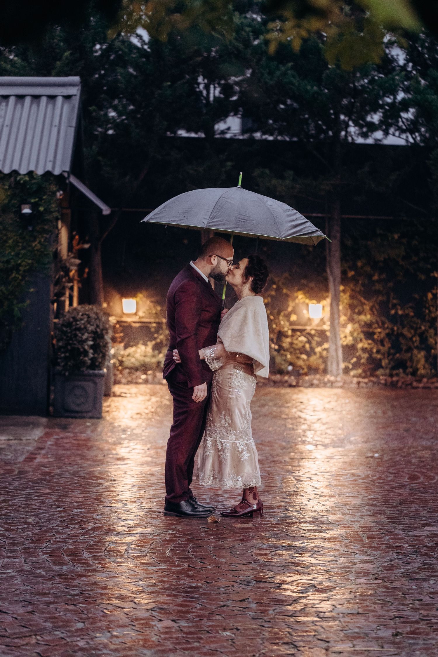 Couple kissing under umbrella in rain on cobblestone path, groom in burgundy suit, bride in lace dress with white wrap.