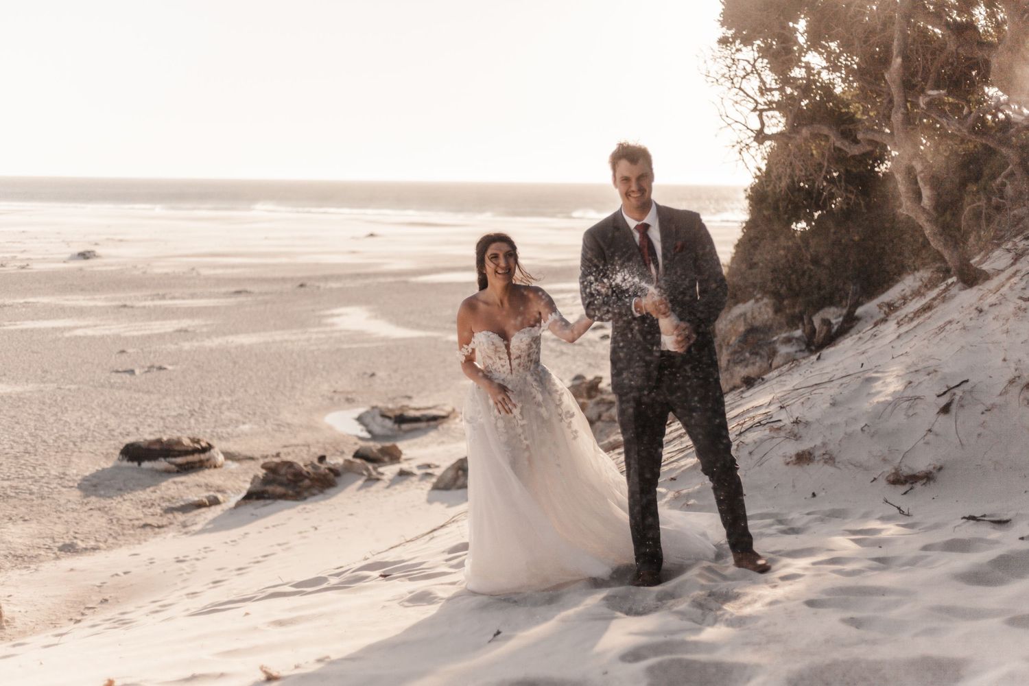 Bride and groom walking on sandy beach dunes at golden hour, bride in lace gown, groom in dark suit.