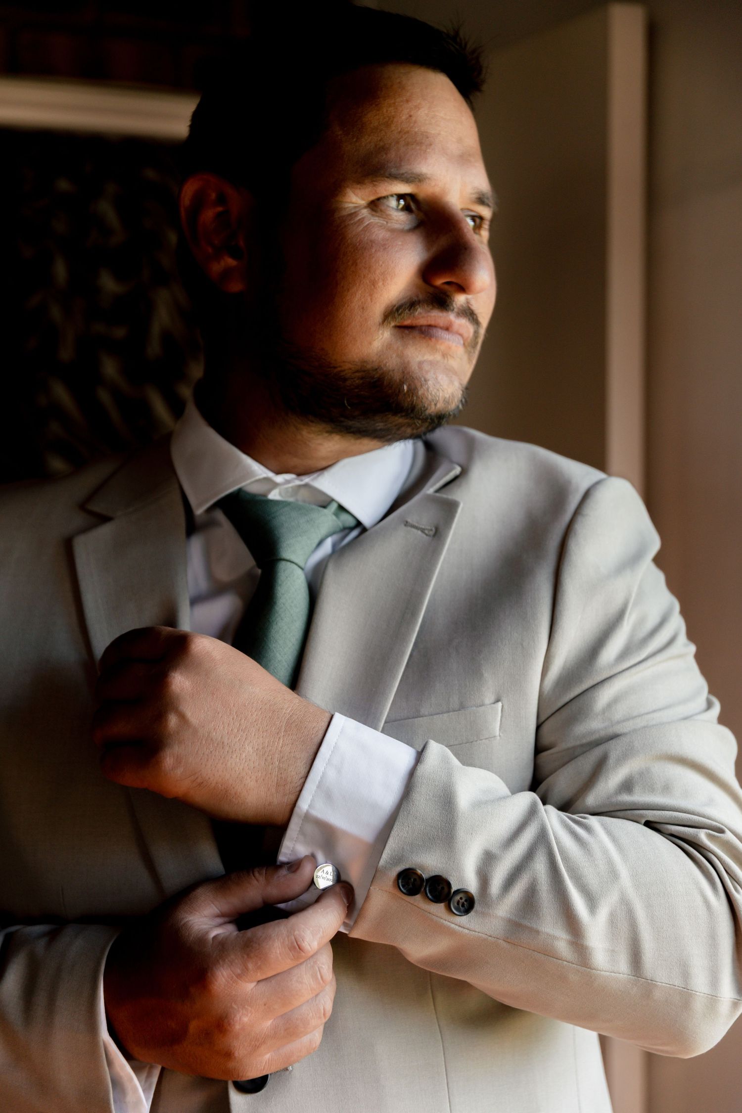 Man in light gray suit adjusting cufflinks, wearing green tie, standing in soft natural light.