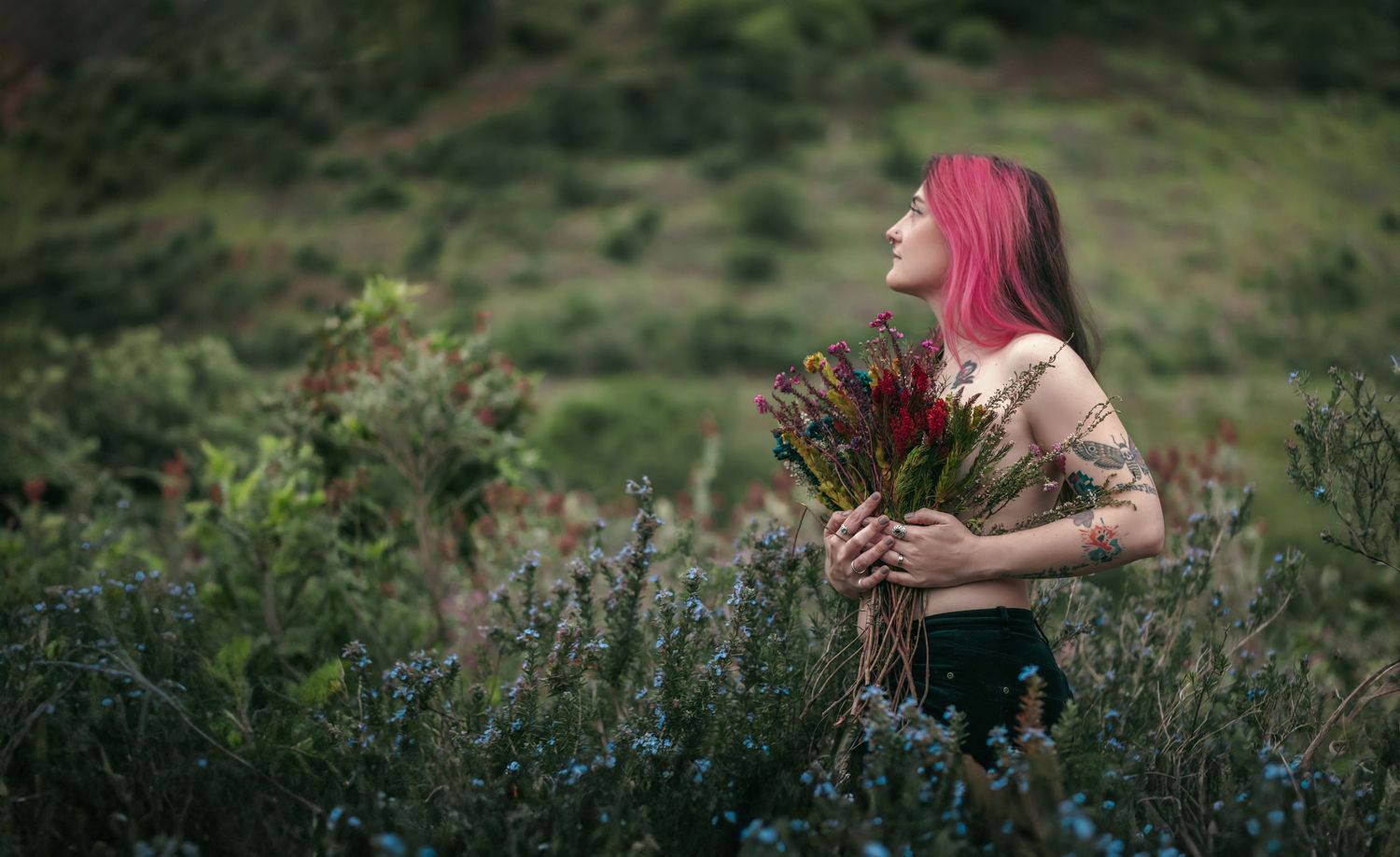 A person with vibrant pink hair stands in a lush green meadow surrounded by wildflowers and natural foliage.