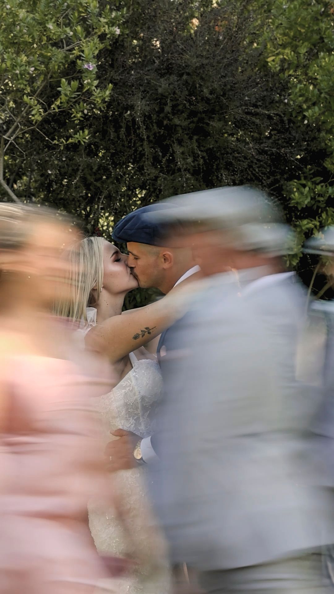 A blurred motion wedding photograph shows a couple sharing a kiss outdoors with green foliage in the background.