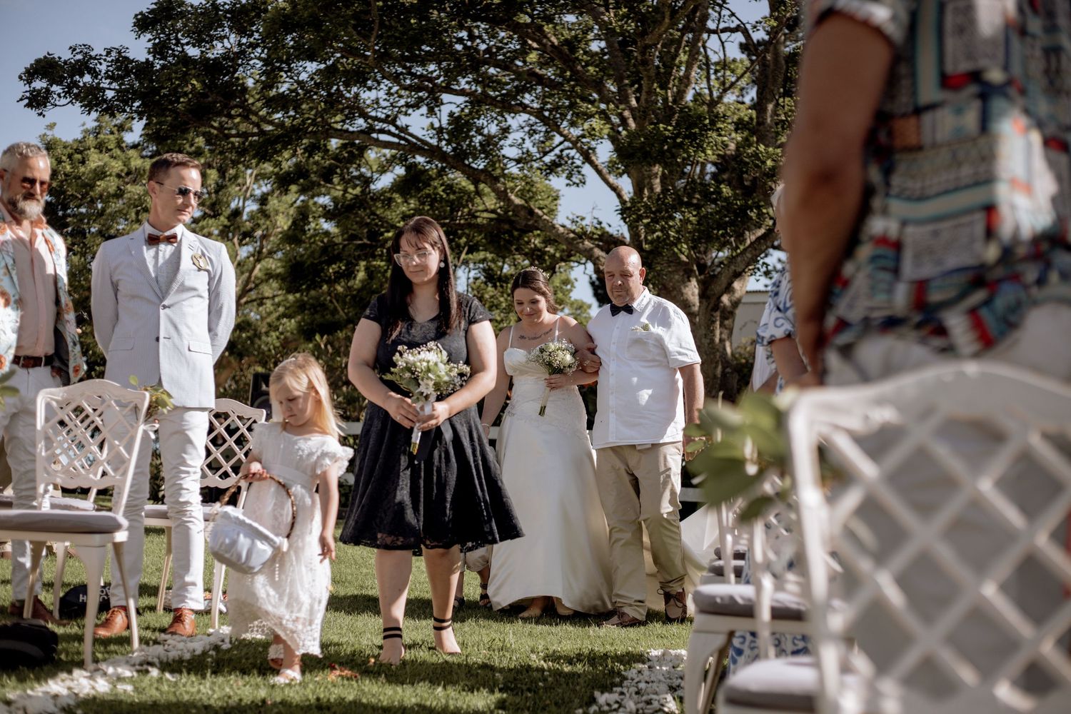 A garden wedding ceremony takes place outdoors with guests seated in white chairs behind a decorative white lattice fence.