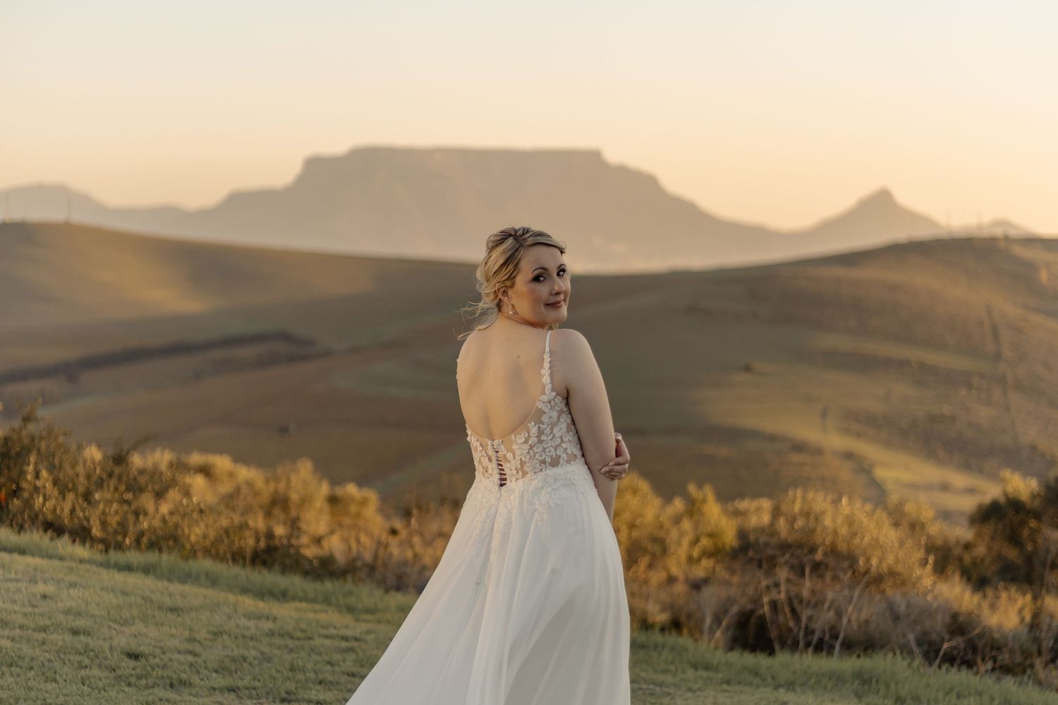 A bride in a flowing white dress stands on a hillside with a majestic mountain silhouetted against the golden sunset.
