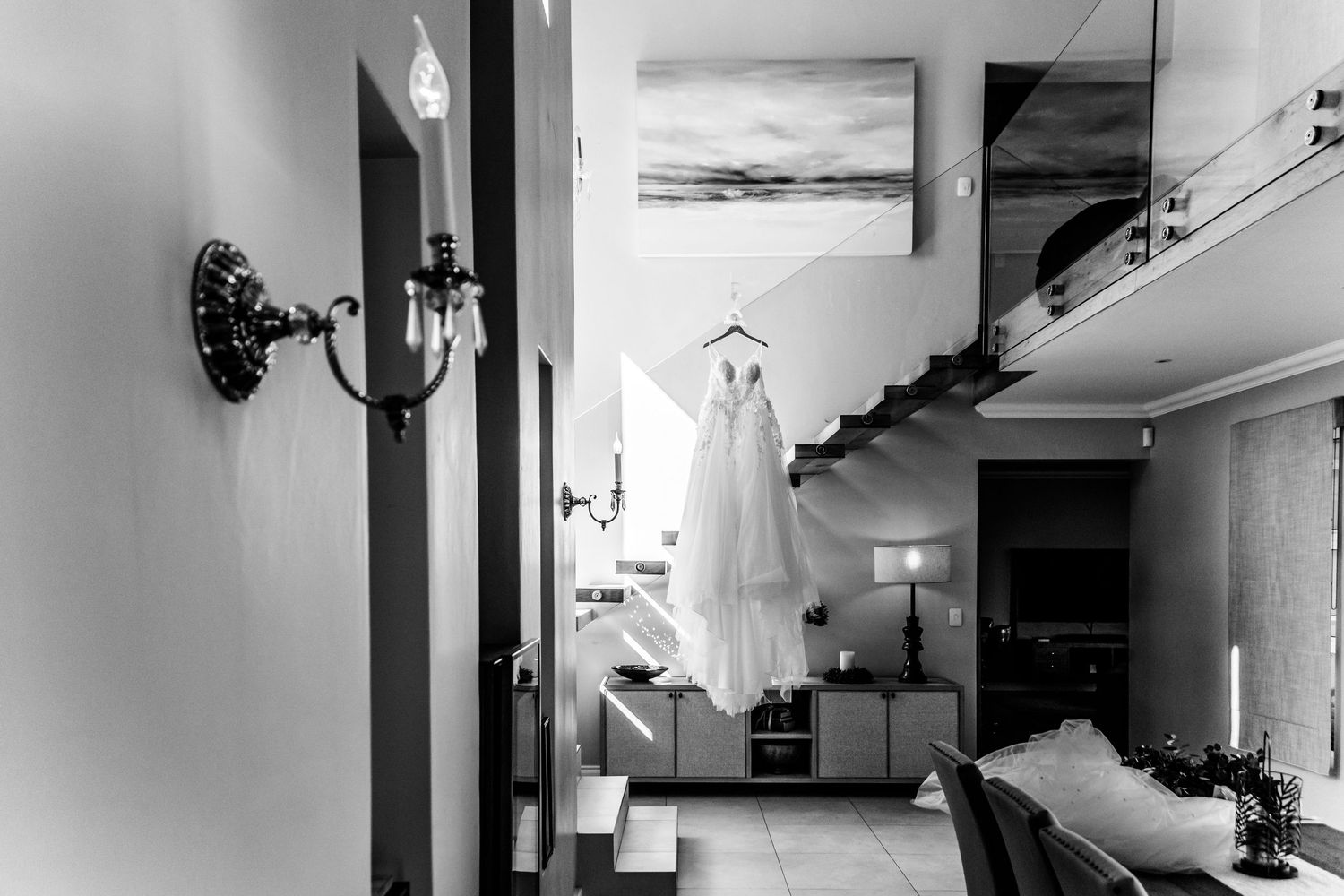 A white wedding dress hangs elegantly in a contemporary interior space with a wall sconce and coastal artwork visible.