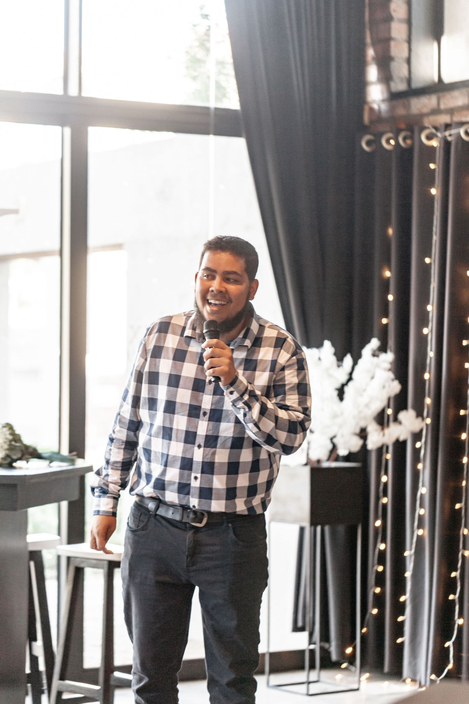 Someone speaking into microphone wearing plaid button-down shirt and jeans in modern space with dark curtains and window.