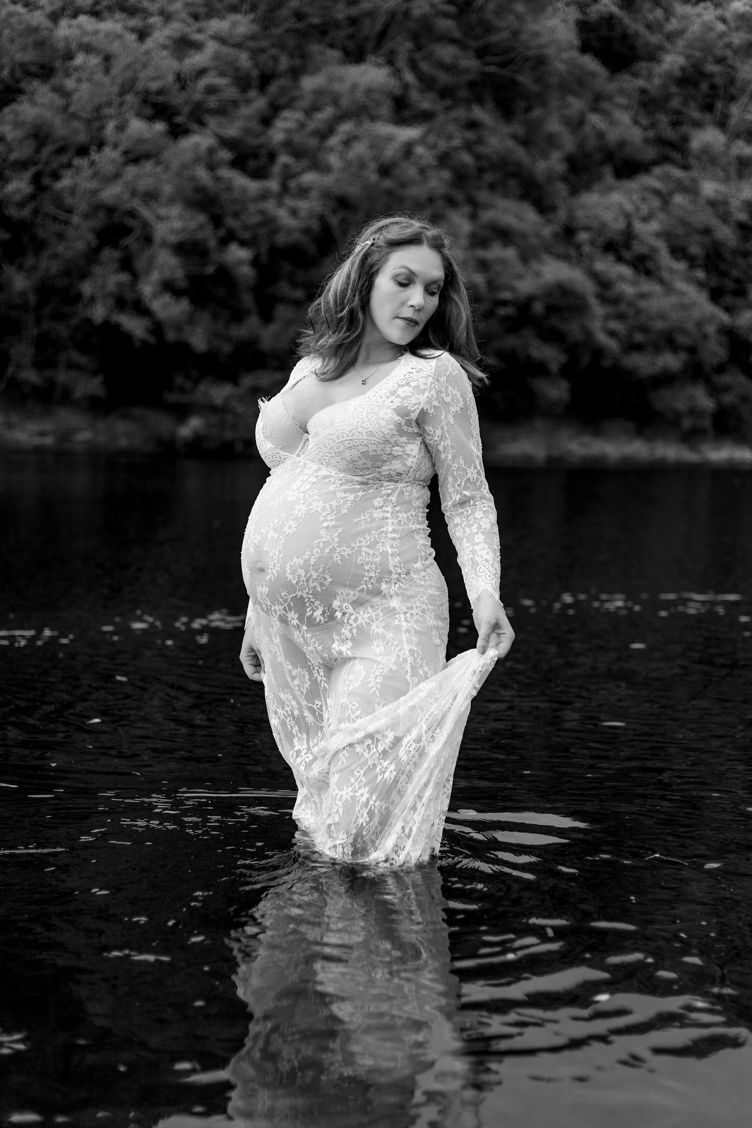A figure in a flowing white dress poses ethereally in dark water against a wooded backdrop, creating a moody artistic portrait.