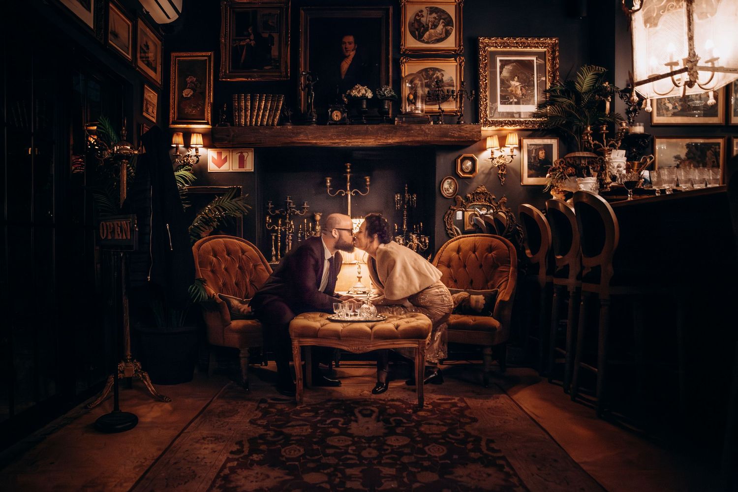 A man sits in an ornate Victorian parlor with antique furniture, candlelight, and portrait-lined walls above a fireplace.