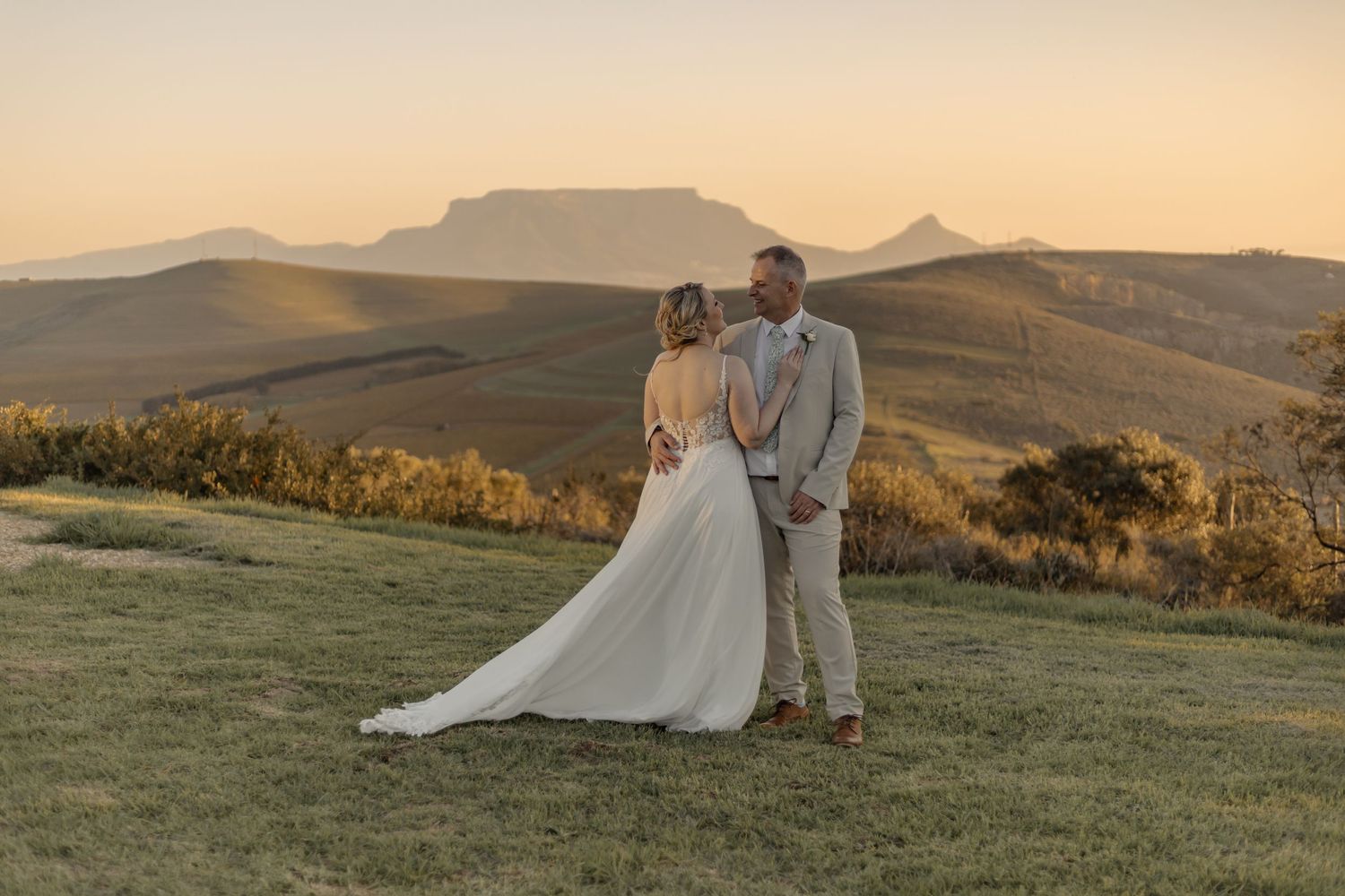 A couple in wedding attire stands on a grassy hill at sunset with mountains silhouetted in the background.
