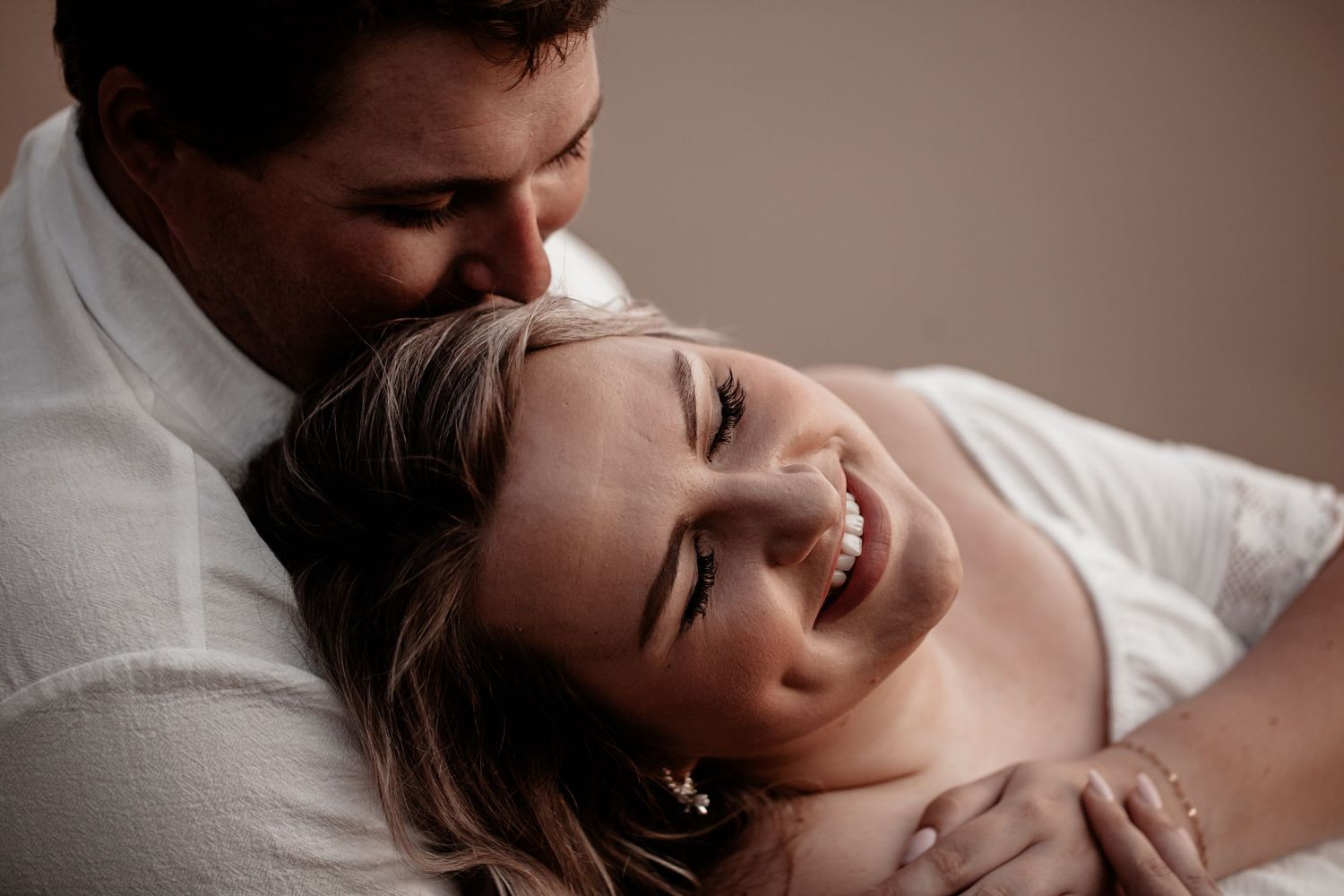 Intimate couple sharing a tender moment together while laughing and embracing on white bedding.