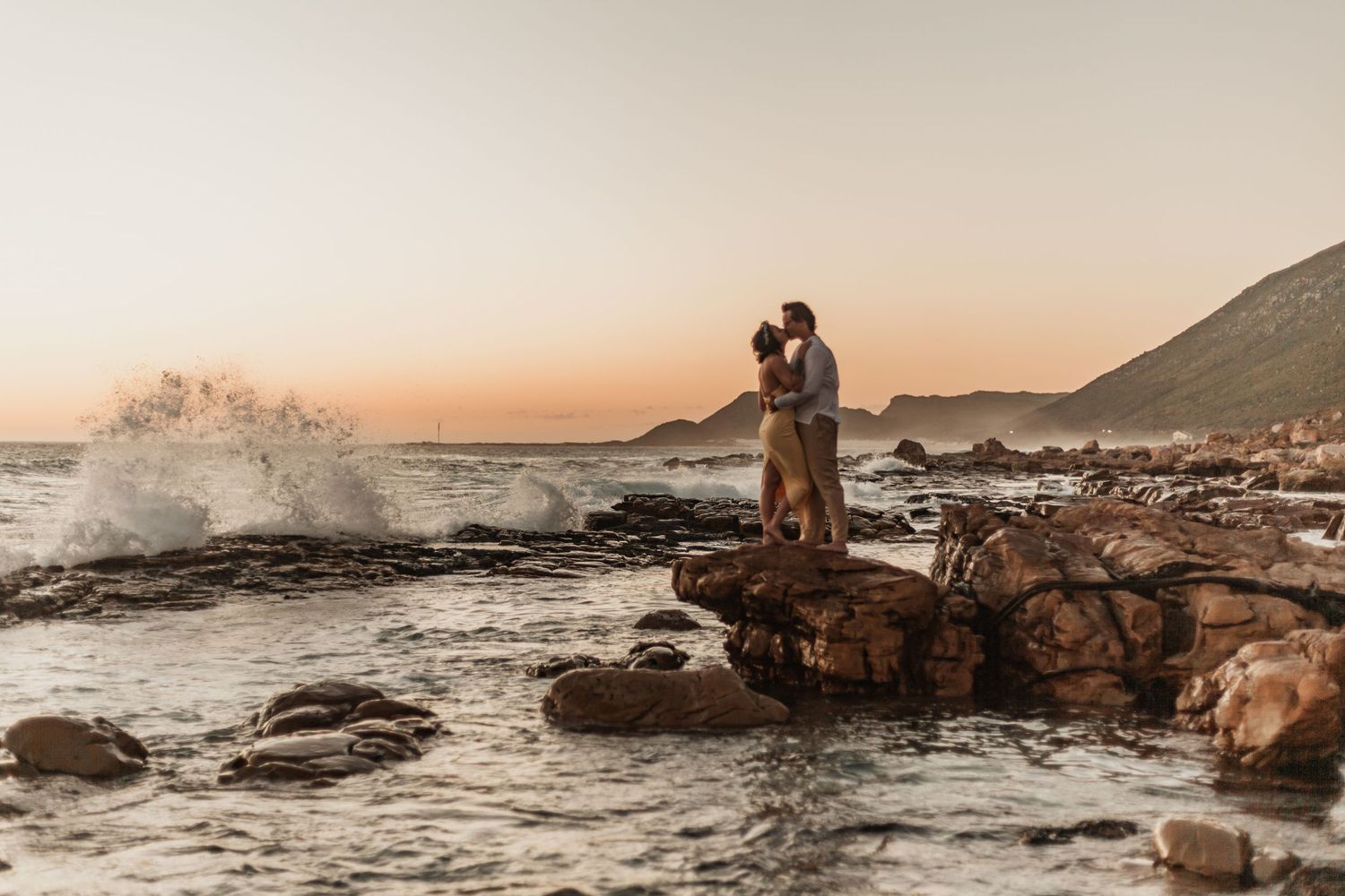 A couple embraces on rocky coastal outcrop at sunset while waves crash against the shore and mountains loom in background.