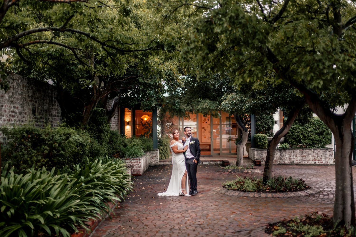 Bride and groom embrace on brick courtyard surrounded by lush trees and stone walls at elegant outdoor wedding venue.