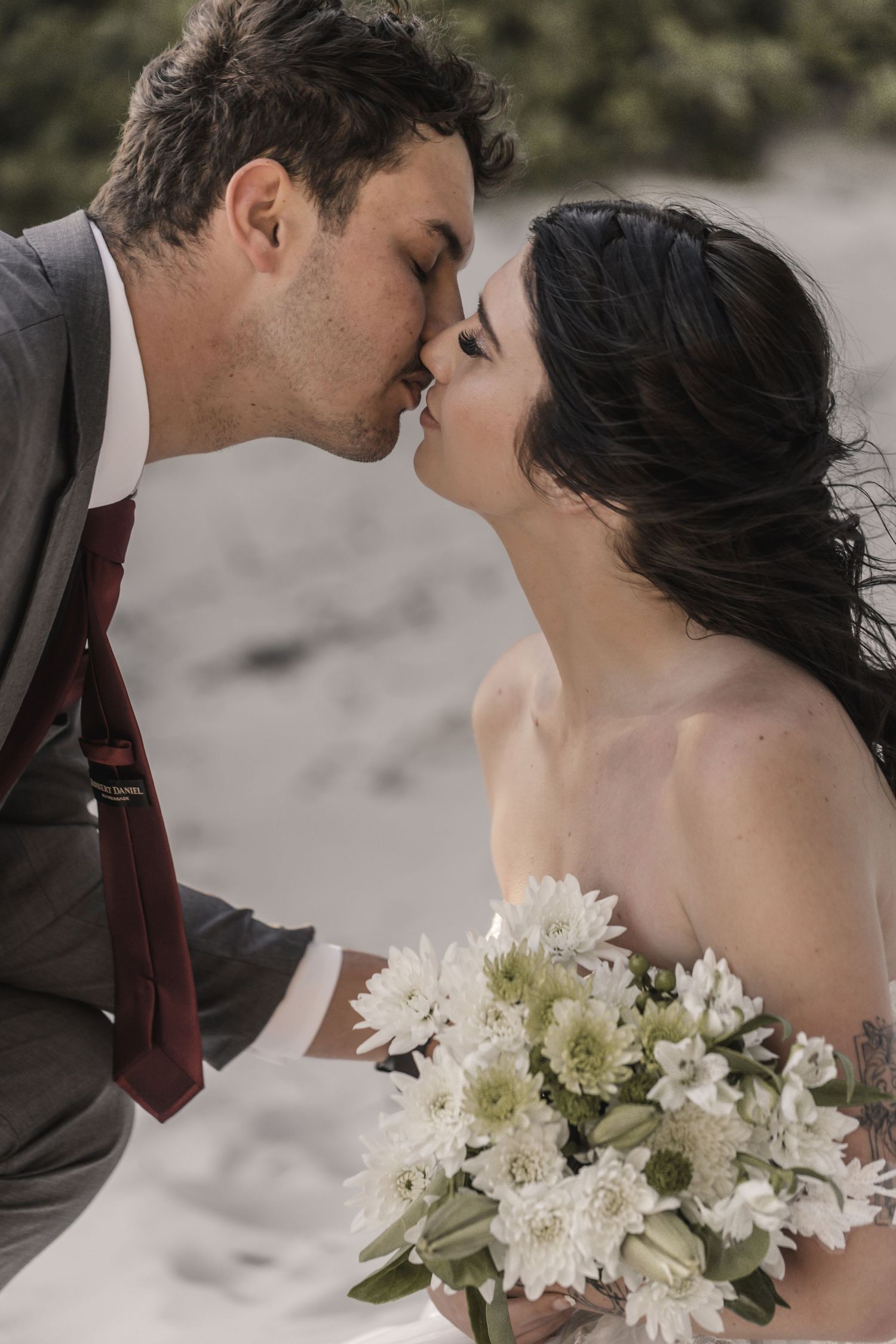 A bride and groom share a romantic kiss on a beach, with the bride holding a bouquet of white flowers.