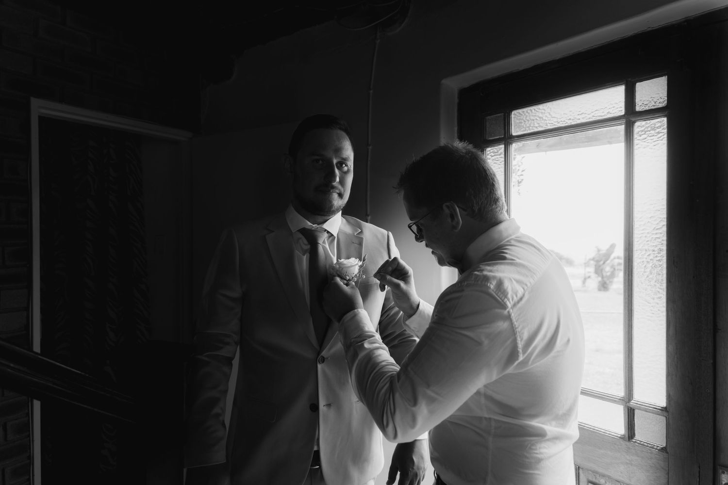 Black and white photo of groom getting boutonniere pinned by groomsman near bright window.