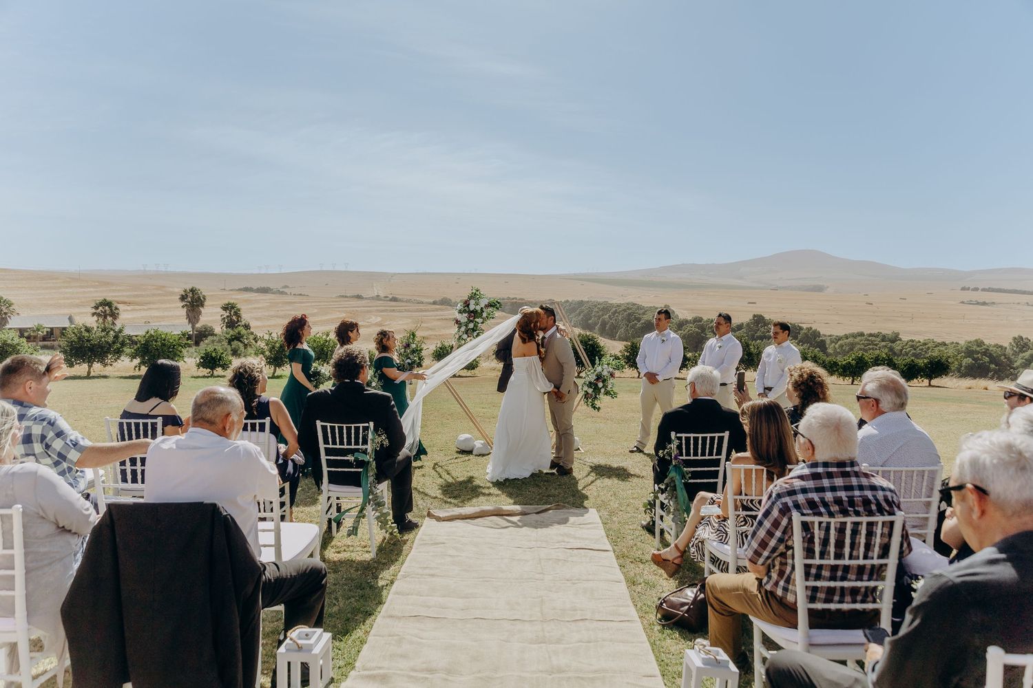 Outdoor wedding ceremony with couple kissing at altar, guests seated on white chairs, scenic golden hills backdrop.