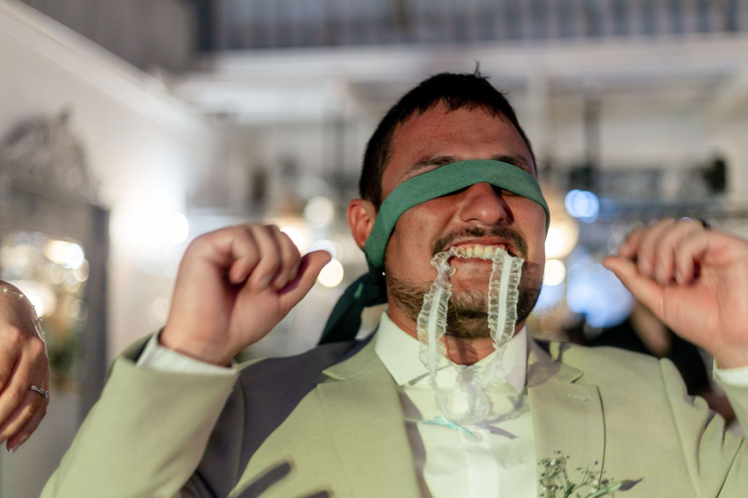 Groom in mint suit blindfolded with green ribbon pulls dollar bills from his mouth at wedding reception.